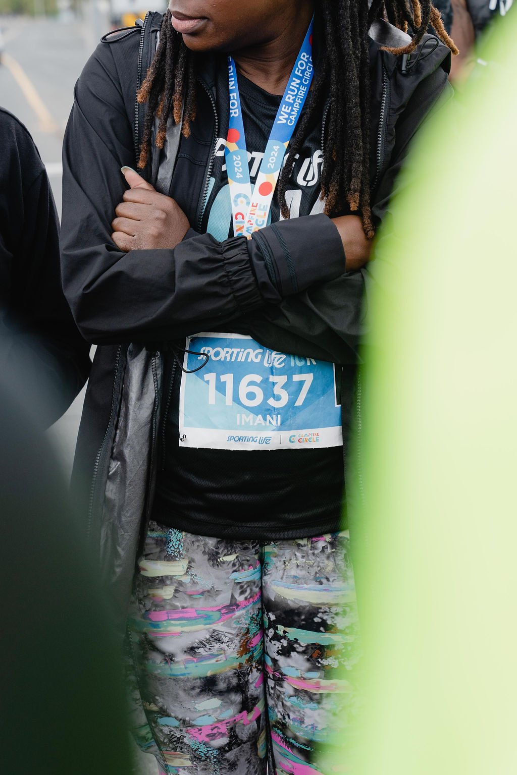 A person with dreadlocks wearing a black jacket, a race bib, and colorful patterned leggings at a marathon event.