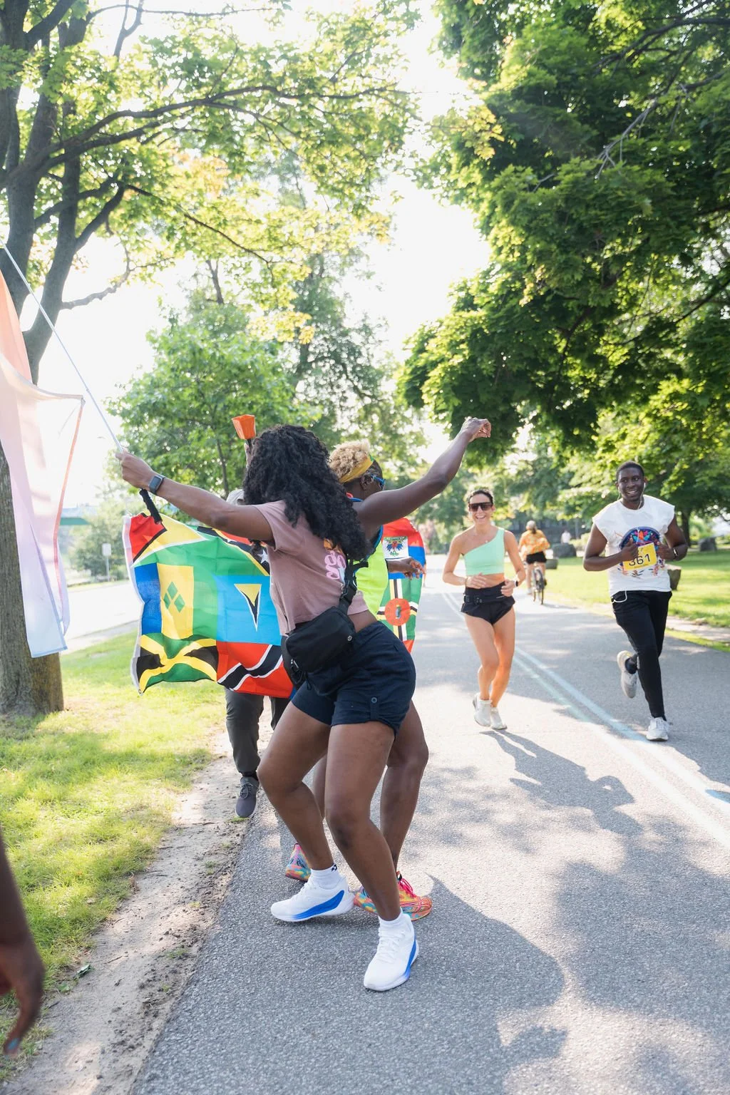 People celebrating at a race event in a park, with a woman dancing near a rainbow flag and runners smiling in the background.