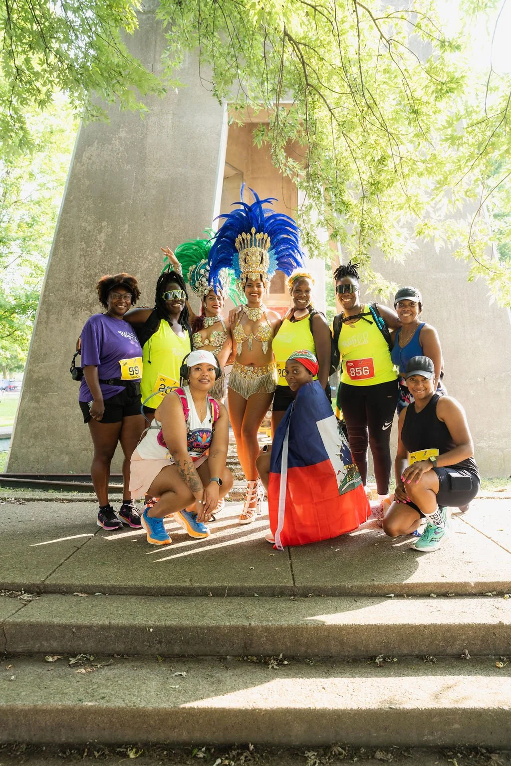Group of women, some wearing bright yellow and purple athletic clothing, and a woman dressed in a traditional carnival costume with a blue feathered headdress, posing outdoors on steps with trees overhead, holding a flag and celebrating.