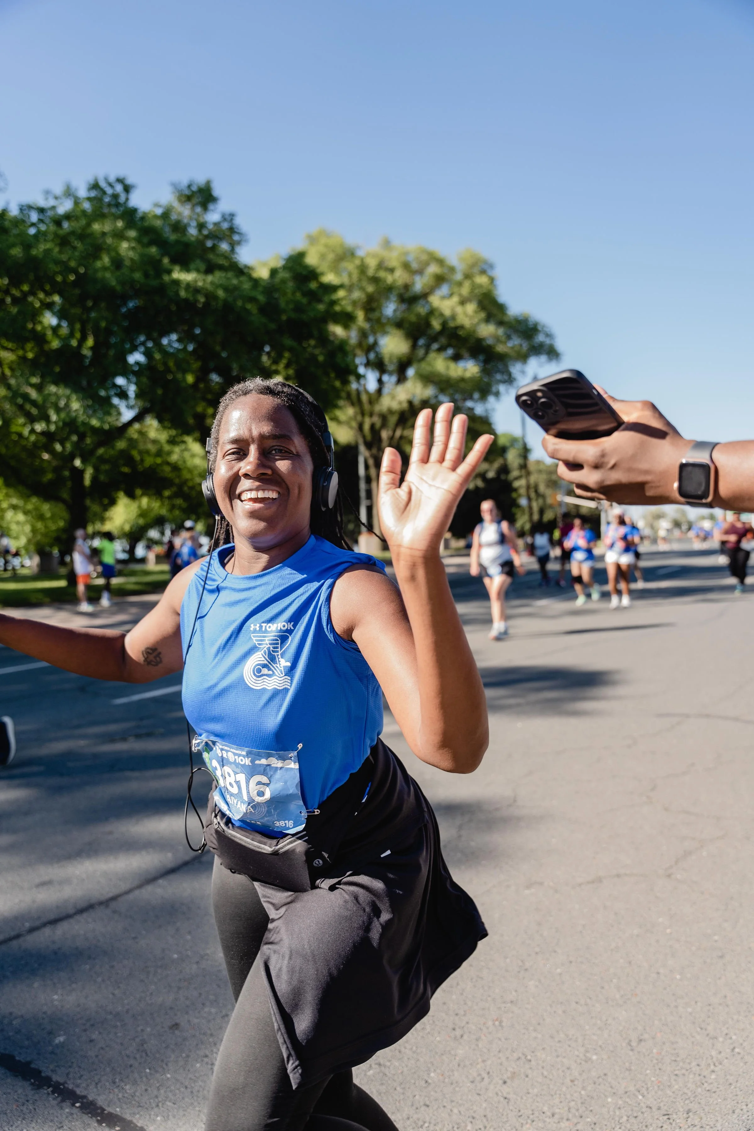 A smiling woman running in a race, wearing a blue athletic shirt and headphones, waving at a person taking her photo against a backdrop of green trees and a clear blue sky.