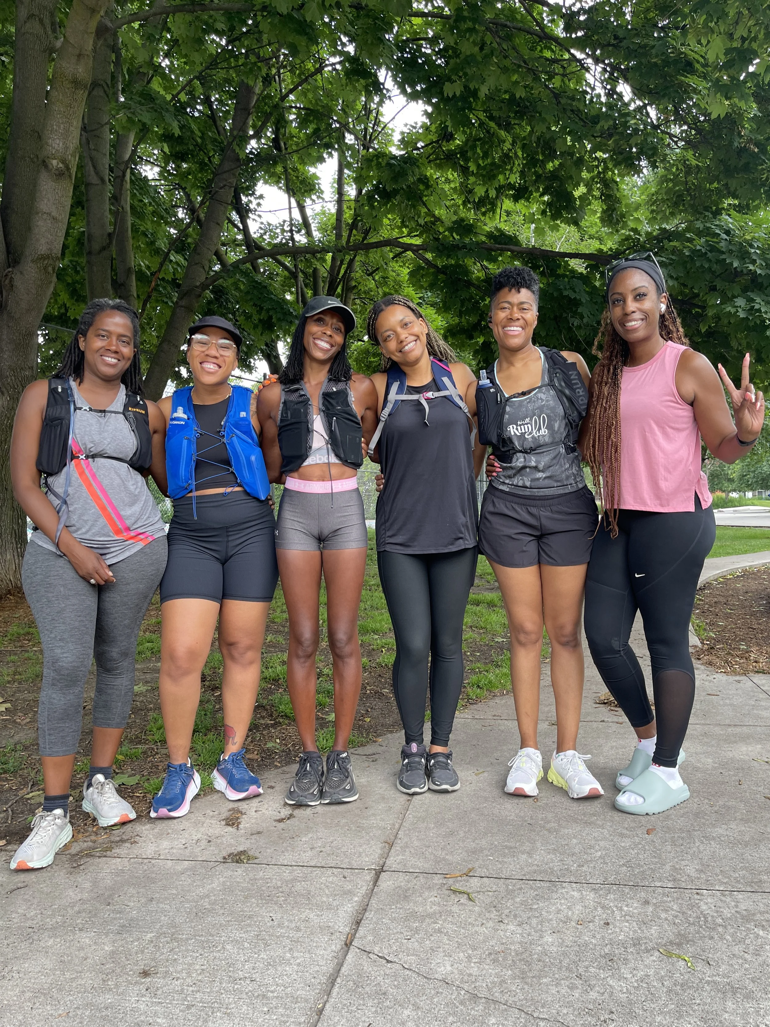 Six women standing side by side outdoors under green trees, smiling at the camera, dressed in athletic wear and carrying backpacks, indicating they are participating in a running or walking event.
