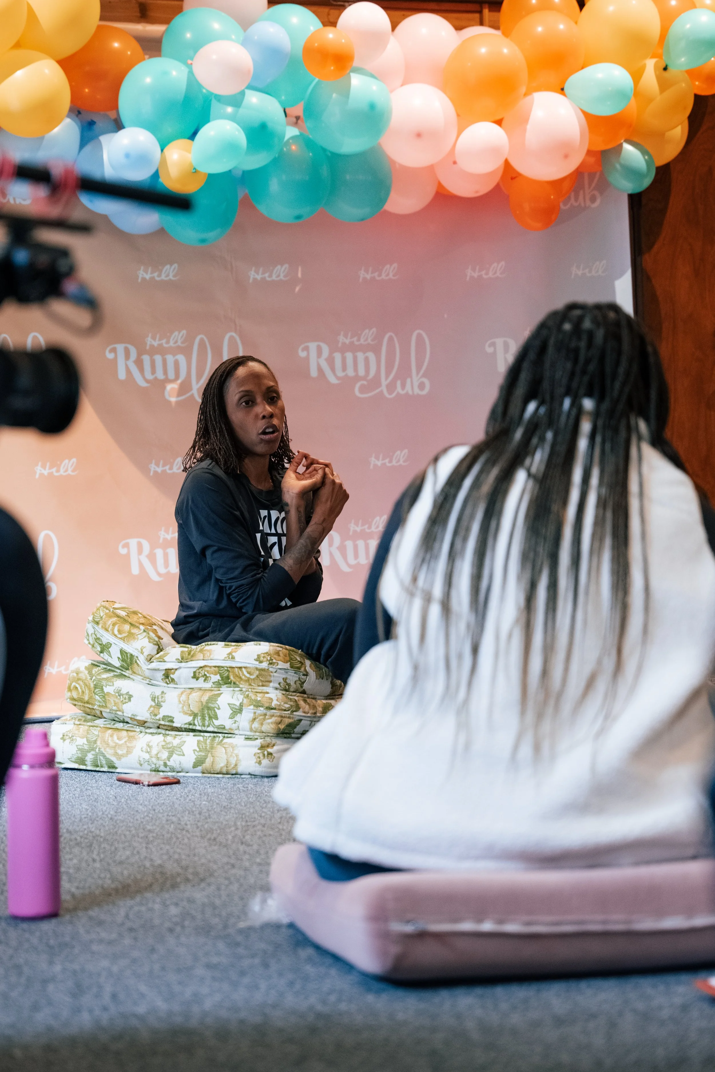 A woman with braided hair sitting on a floral cushion, speaking during a discussion at the Hill Run Club event. The background features a pink wall with balloons in shades of orange, white, yellow, blue, and teal, and the text 'Hill Run Club' is visi