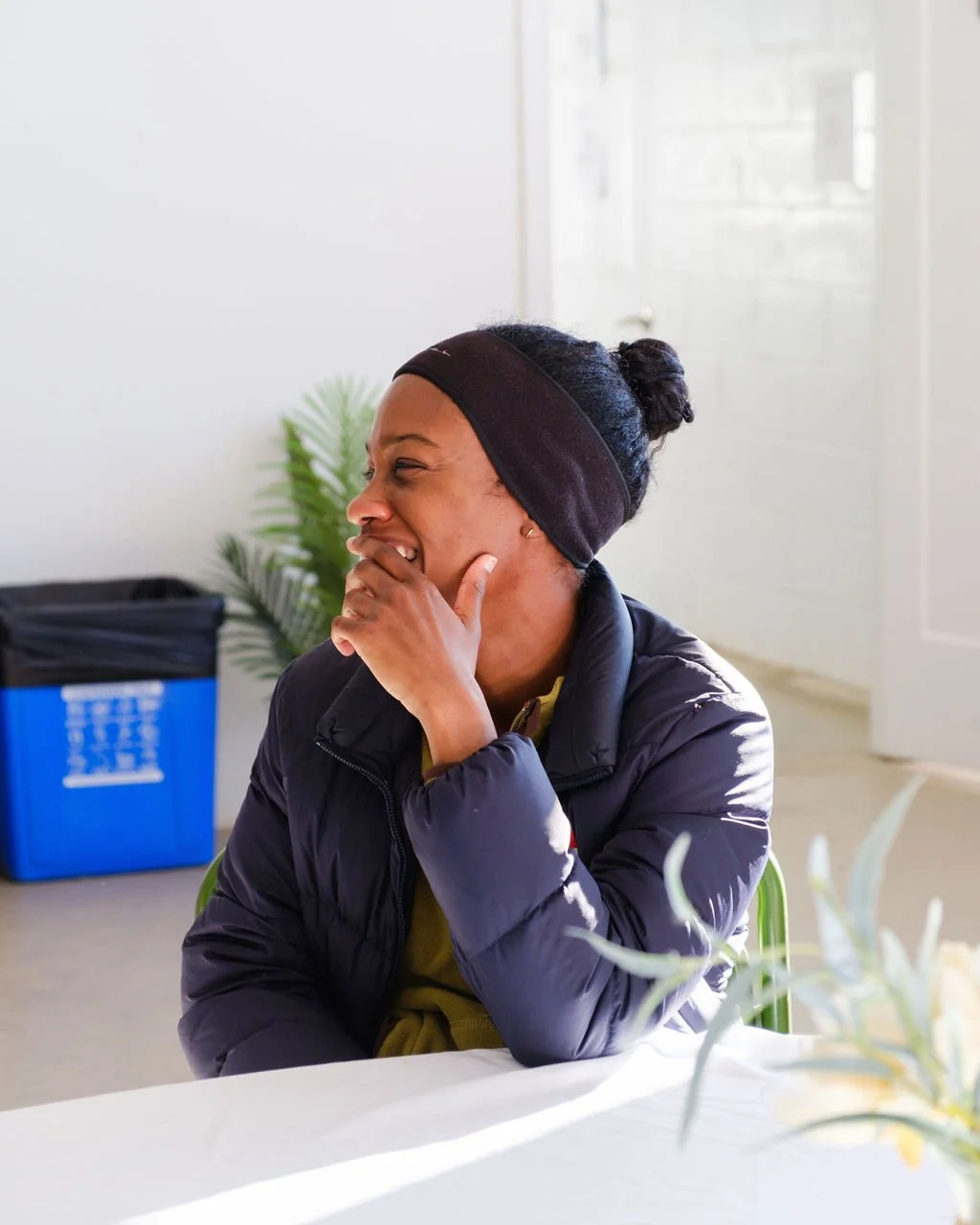 A woman laughing with her hand covering her mouth, seated indoors at a table with a white tablecloth, wearing a black headband and a dark jacket.