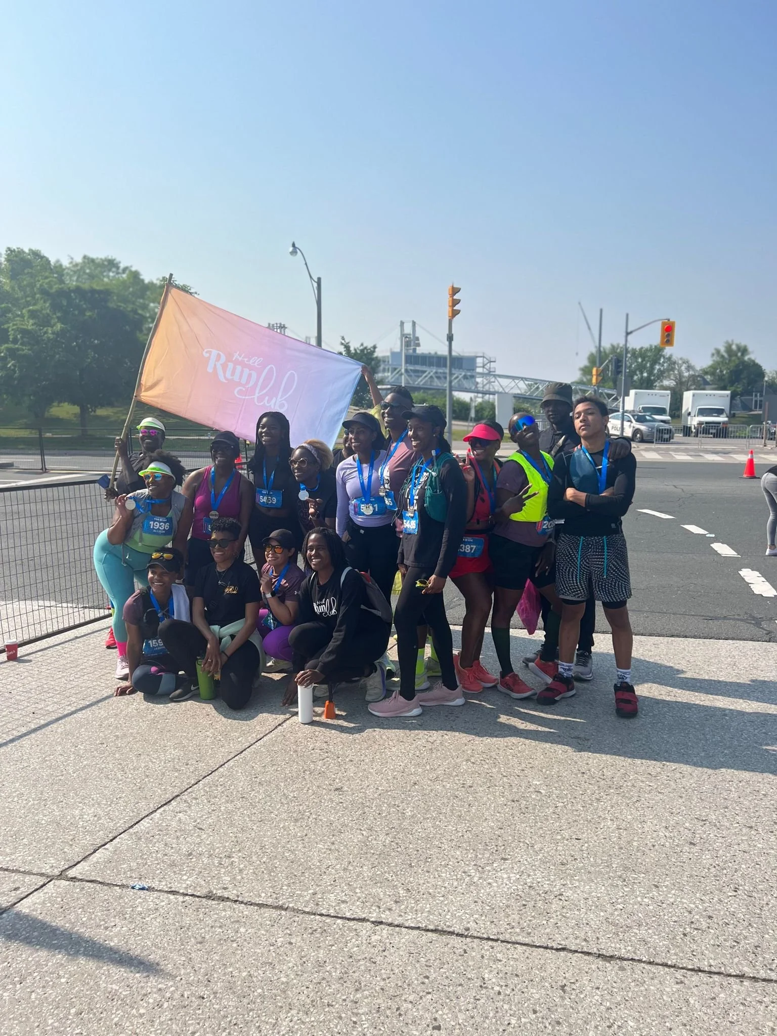 Hill Run Club runners at a race event holding a pink flag that says 'Hill Run Club,' with some wearing medals and athletic gear, standing on a sidewalk near a street with traffic lights and trucks in the background.