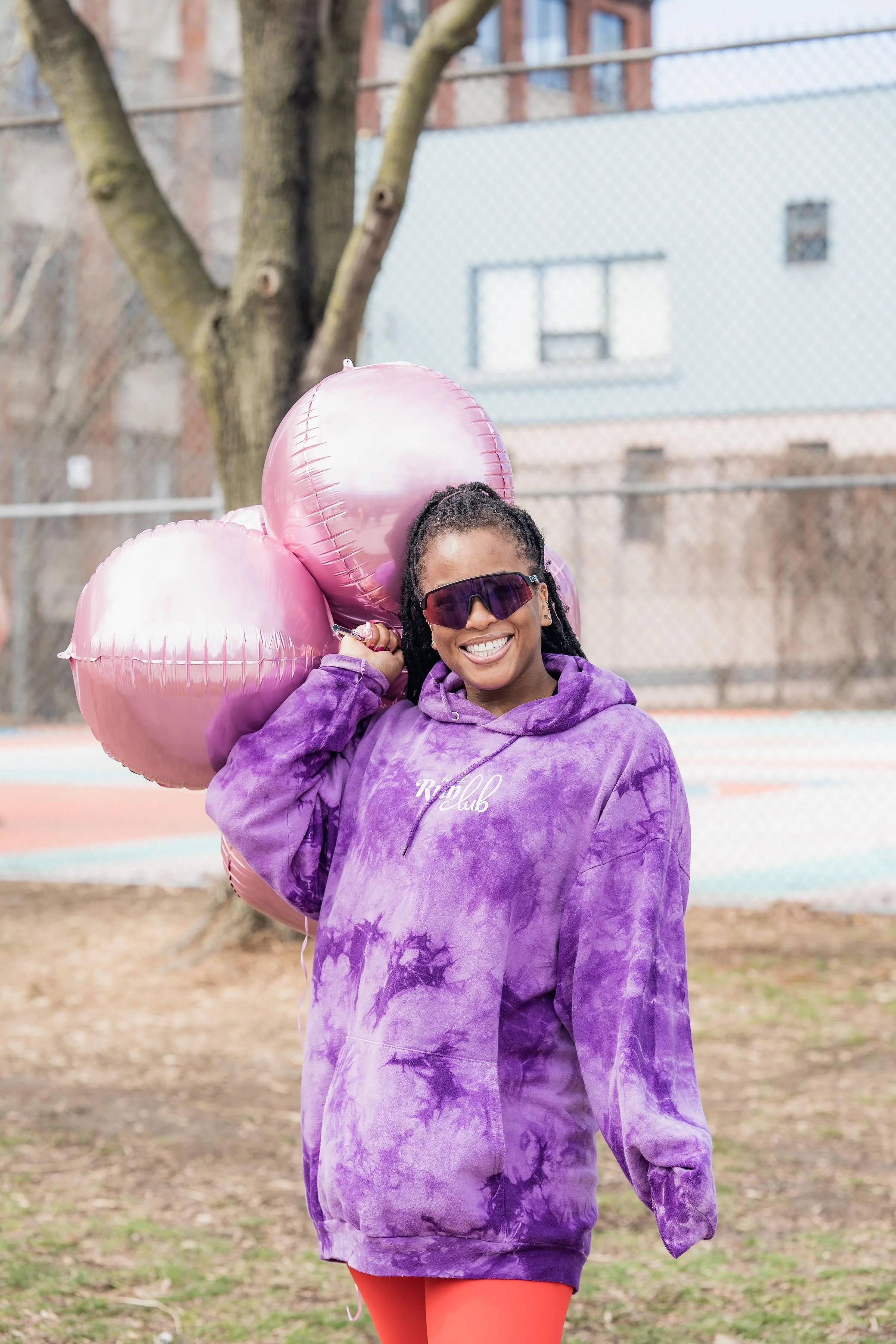 A woman with dreadlocks, wearing sunglasses and a purple tie-dye hoodie that says 'KIP Club,' holding three pink heart-shaped balloons, standing outdoors on a park or playground with a tree and a chain-link fence in the background.