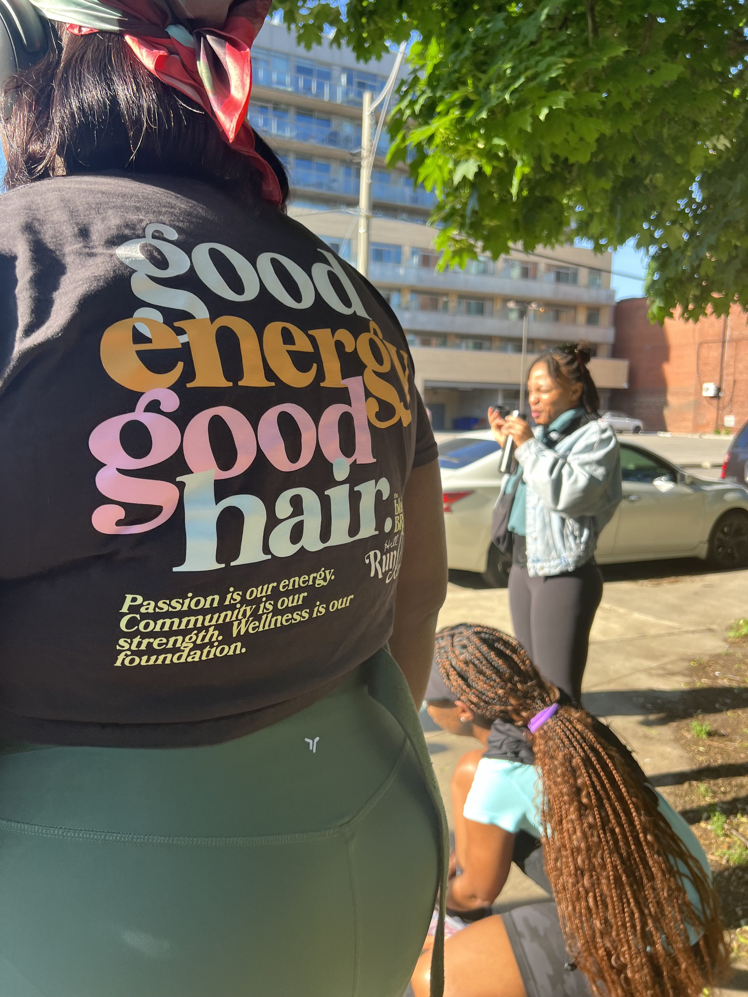 A woman with hooked hair sitting on the sidewalk using her phone, a woman with long braided hair and a purple hair tie sitting nearby, and another woman standing and looking at her phone, outdoors with trees and buildings in the background. The foreg
