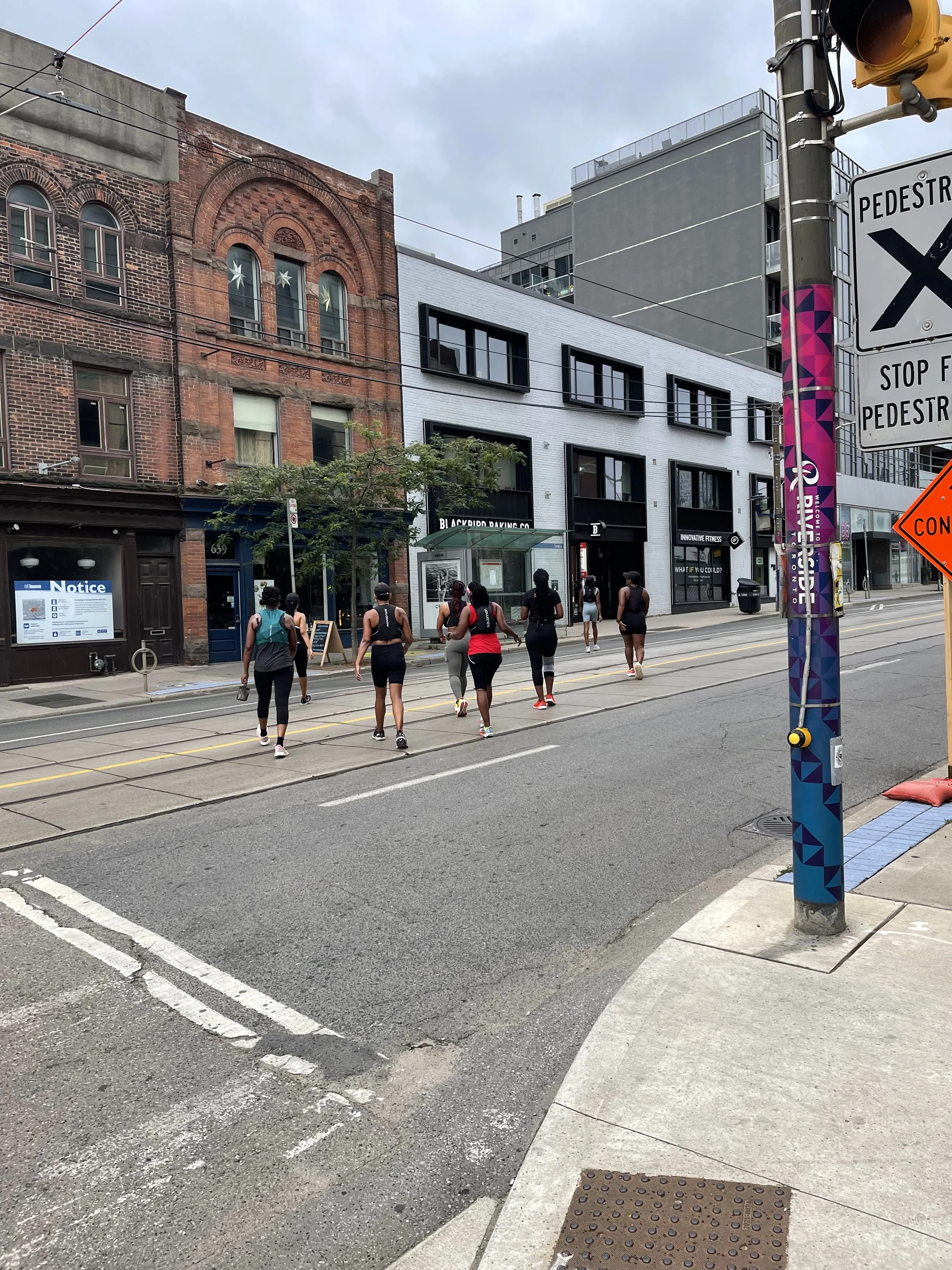 Group of people walking on the street, near a bus stop, with modern and historic buildings in the background on a cloudy day.