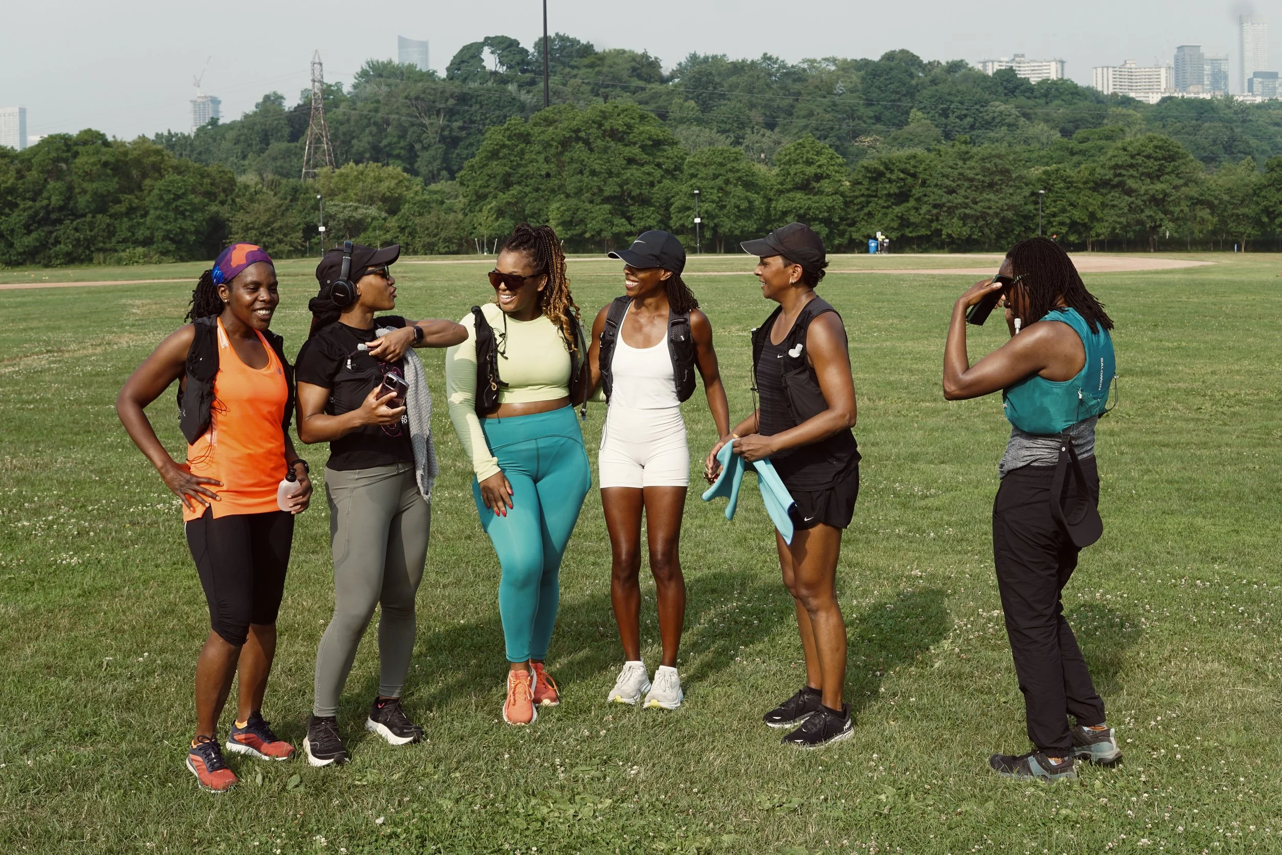Six women in athletic clothing standing on a grassy field, talking and smiling, with trees and city buildings in the background.