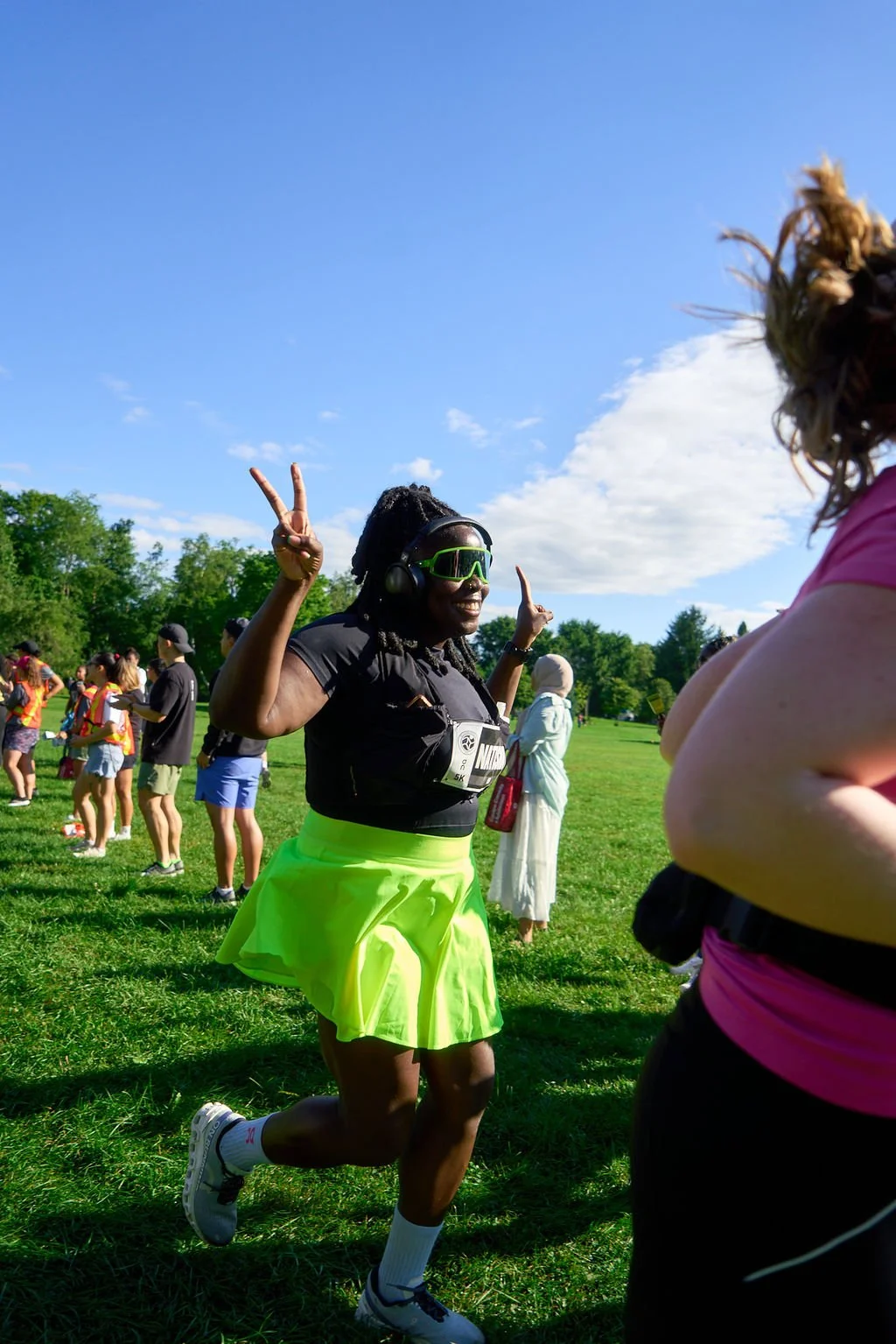 A woman running outdoors during a race, wearing a black t-shirt, bright yellow skirt, white socks, and running shoes, making peace signs with both hands, and smiling under sunny skies with other participants and spectators in the background.