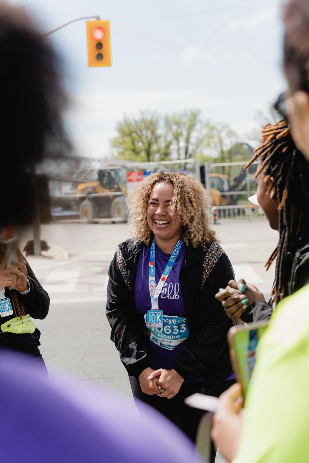 A smiling woman with curly blonde hair wearing a purple race shirt and a race bib, surrounded by a group of people and standing at a road intersection with a traffic light overhead.