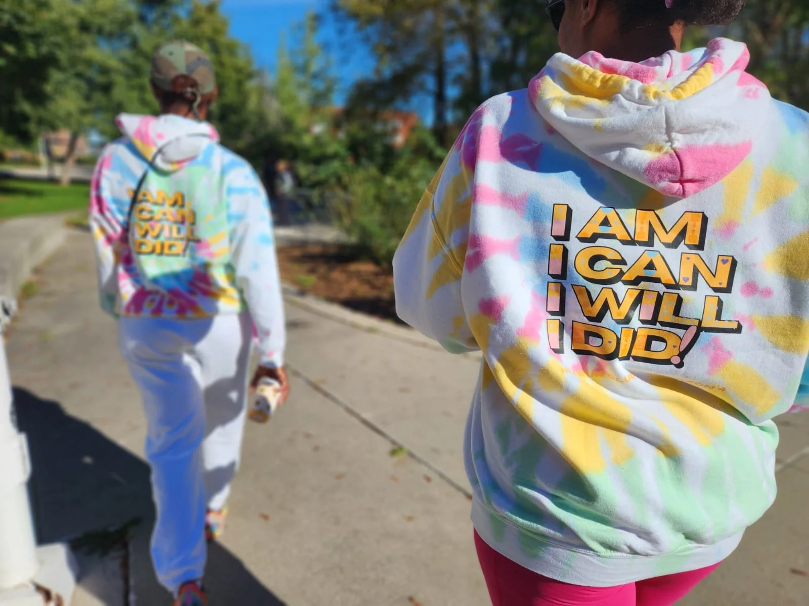 Two people wearing tie-dye hoodies with the phrase 'I AM I CAN I WILL I DID!' printed on the back, walking outdoors on a sidewalk in a park area with trees and a blue sky.