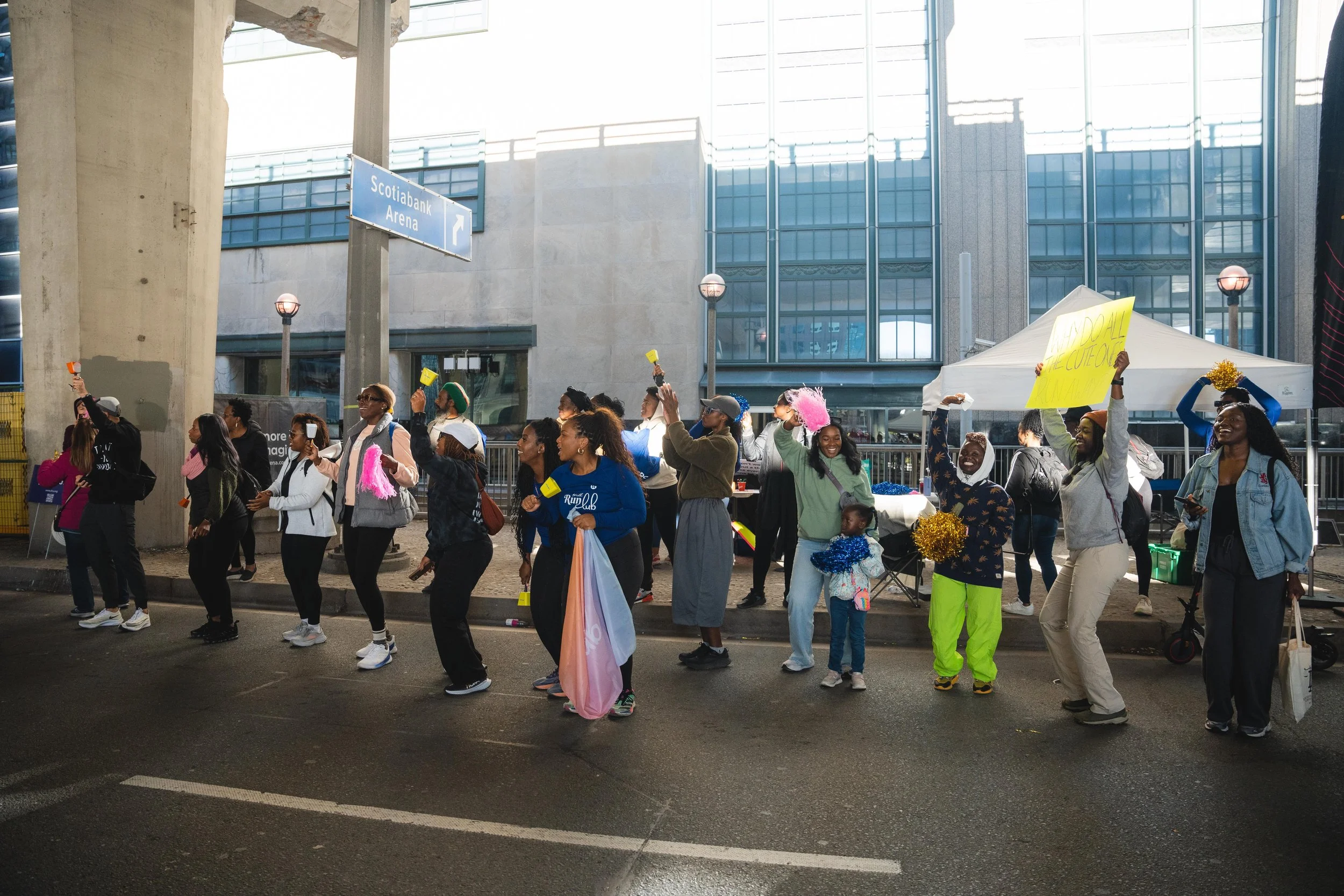 A diverse group of people participating in a protest or rally on a city street, some holding signs and pom-poms, with a tent and city buildings in the background.