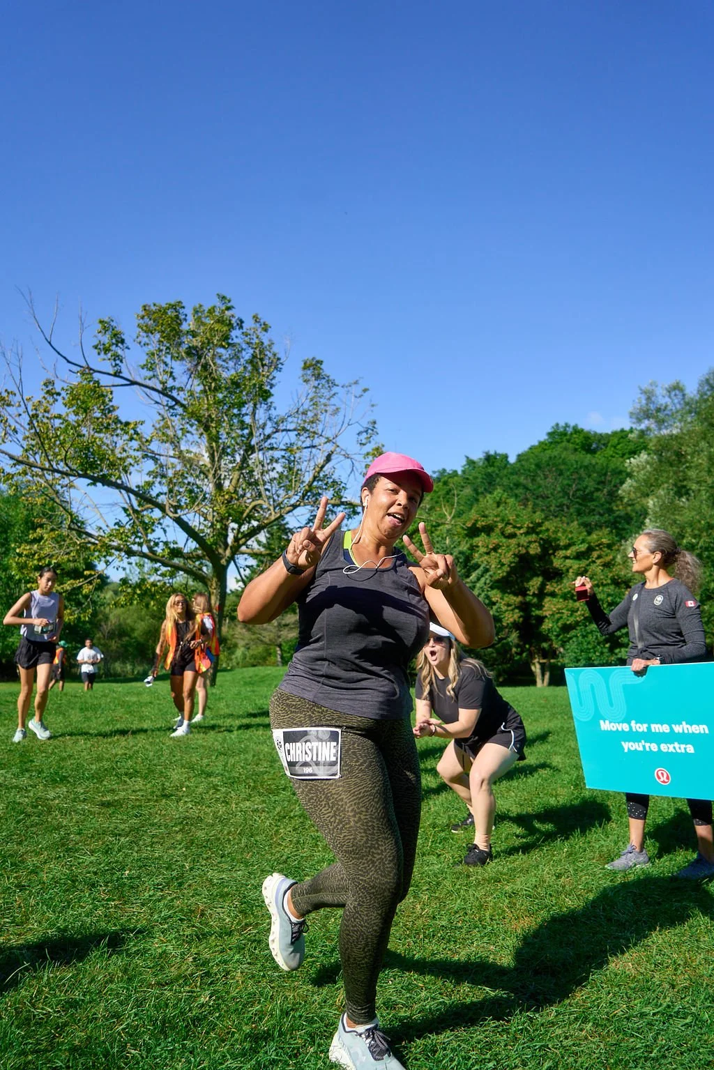 A woman running in a park during a race, making peace signs and smiling, with a group of women and a volunteer holding a sign in the background. The scene is bright and sunny with green trees and a clear blue sky.