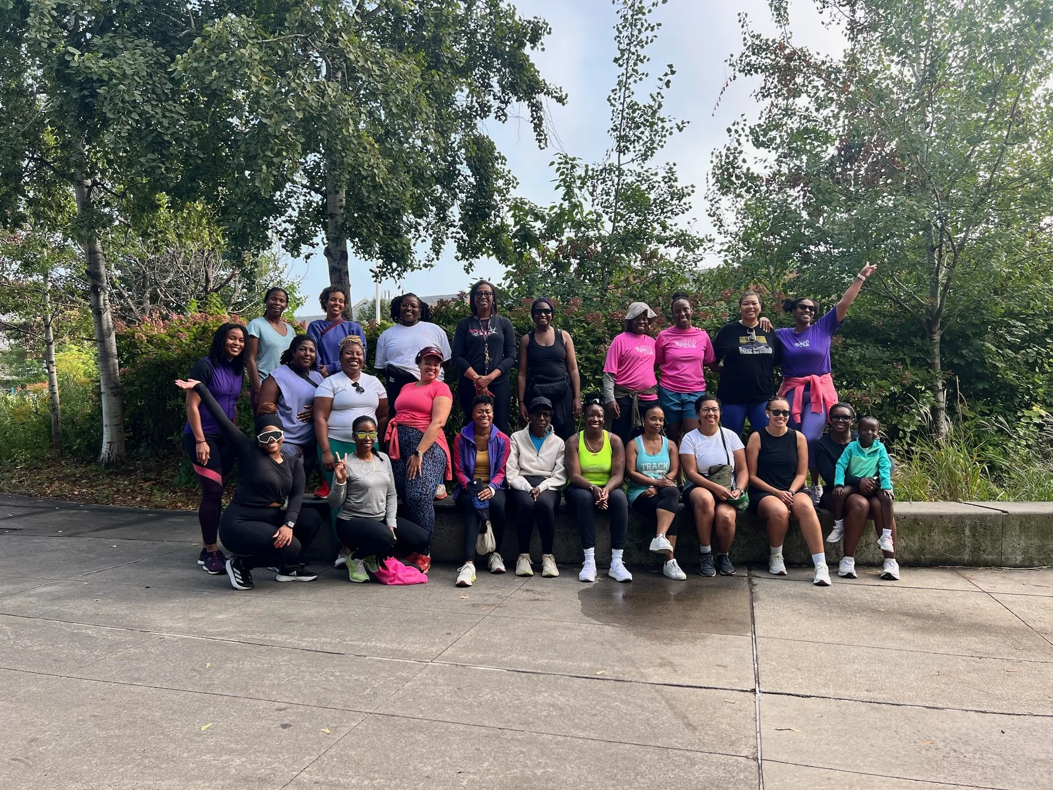 A group of women and girls posing outdoors in front of trees, some sitting and some standing, dressed in athletic and casual clothing.