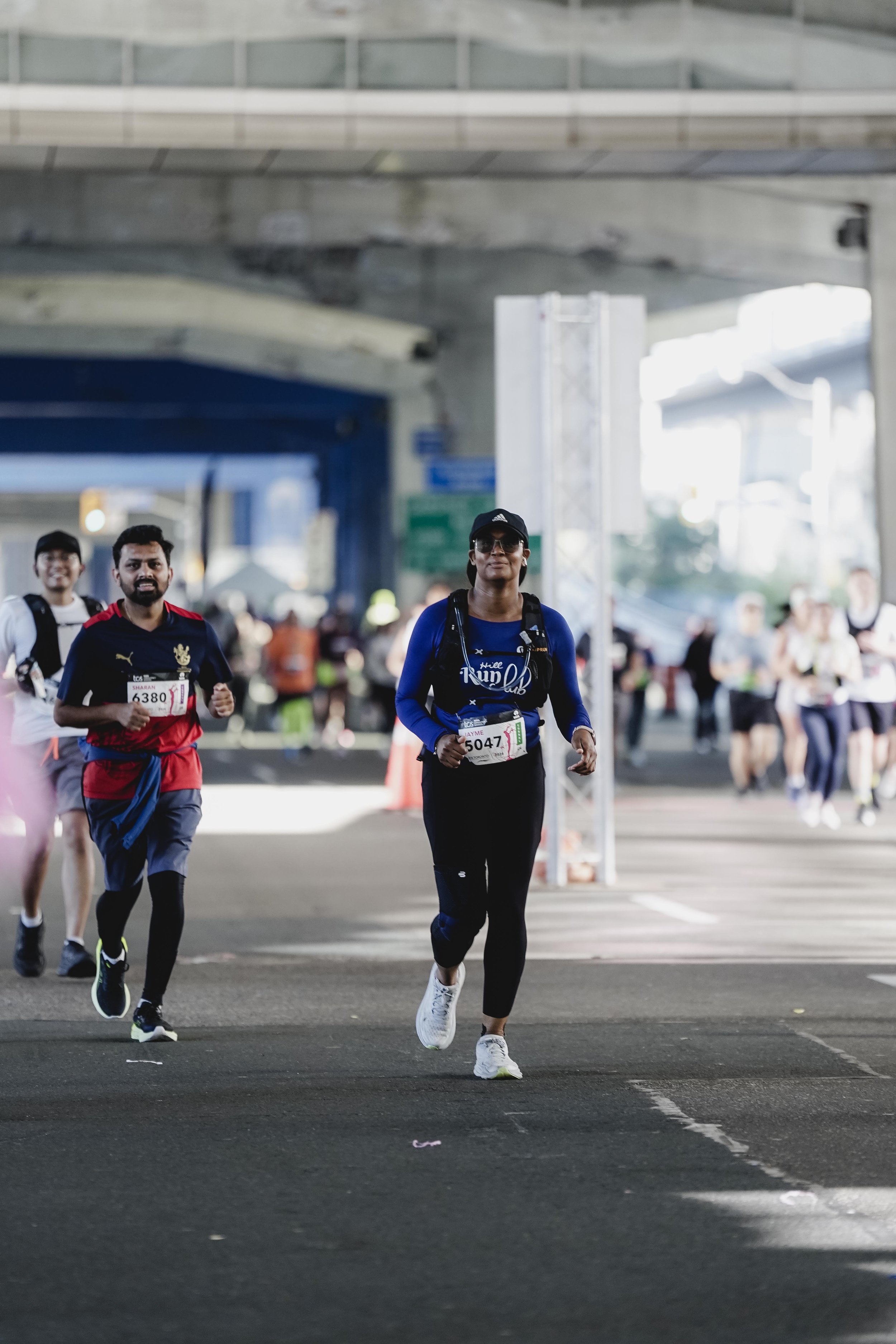 People participating in a marathon under an overpass, with some runners wearing athletic gear, race bibs, and backpacks, in an urban setting.