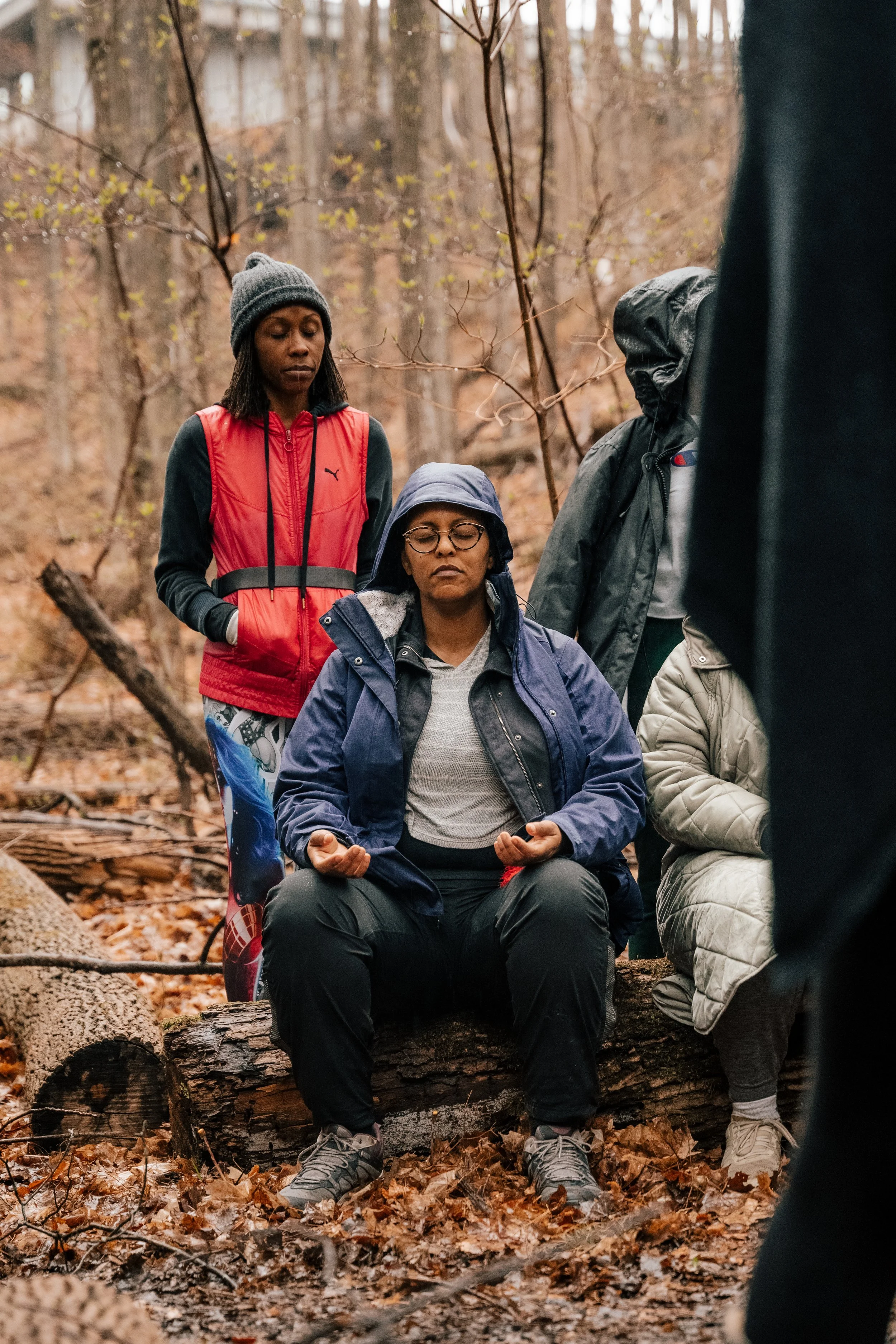 A woman with glasses, wearing a blue jacket and gray hoodie, sits on a log with her eyes closed and hands in a meditative pose, surrounded by other people in outdoor clothing in a wooded area.
