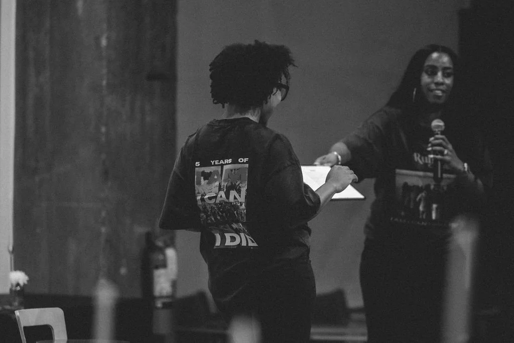 Two women on stage: one speaking into a microphone and the other facing away with a clipboard, in a dark room