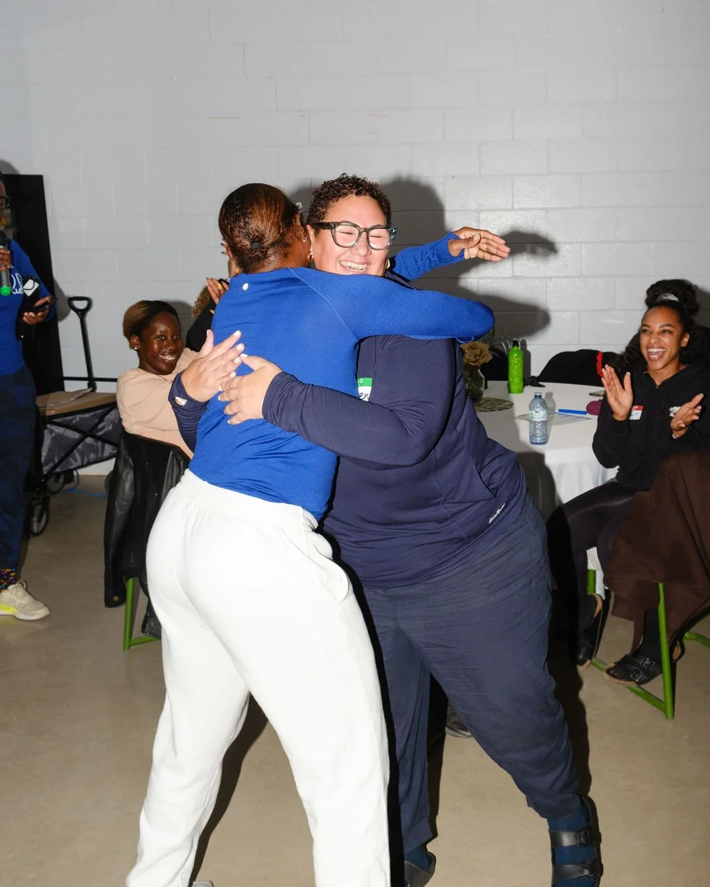 Two women hugging each other with smiles, surrounded by seated women clapping and smiling at a celebration or gathering in an indoor space with tables and chairs.