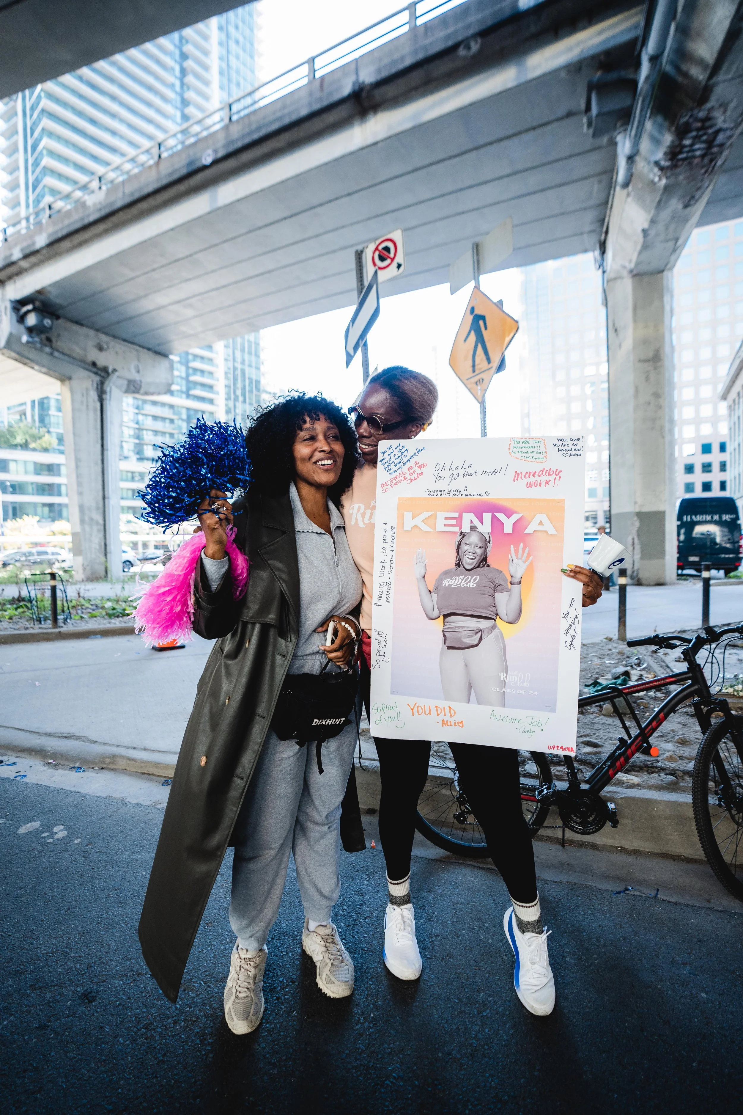 Two women standing under an overpass, smiling and holding a large framed photograph of a woman with the word 'KENYA' at the top. The woman on the left is holding pink and blue pom-poms, while the woman on the right is hugging her. There are bikes, pa