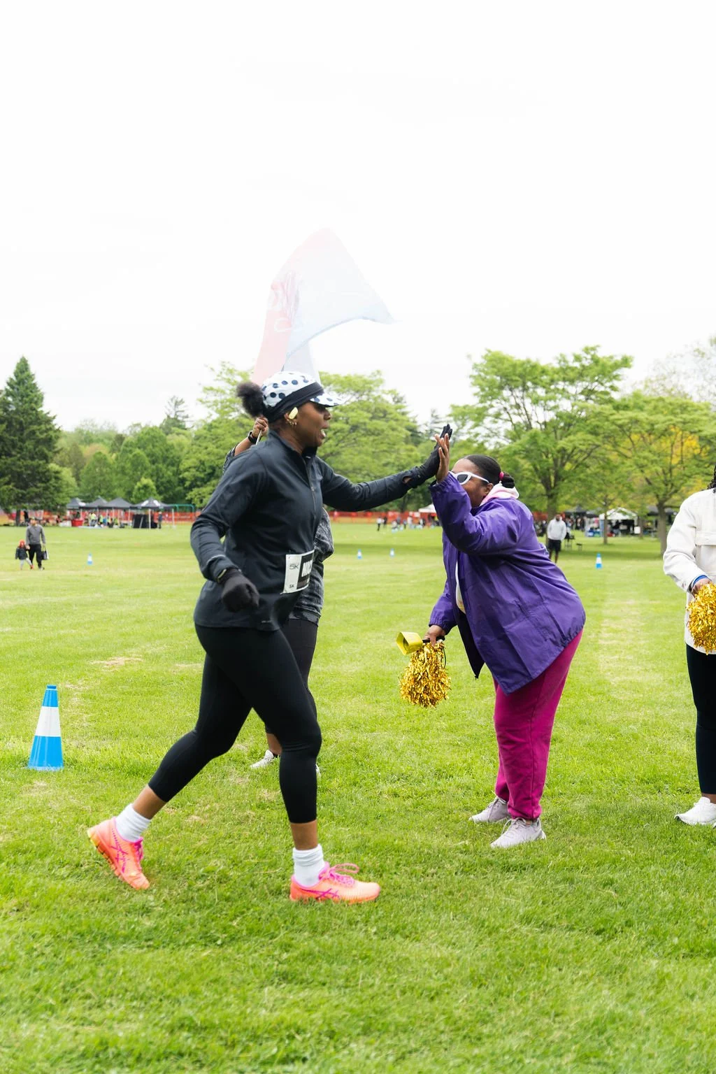 A woman in a black jacket and running gear high-fives a woman in a purple jacket and maroon pants at an outdoor event on a grassy field with trees in the background.