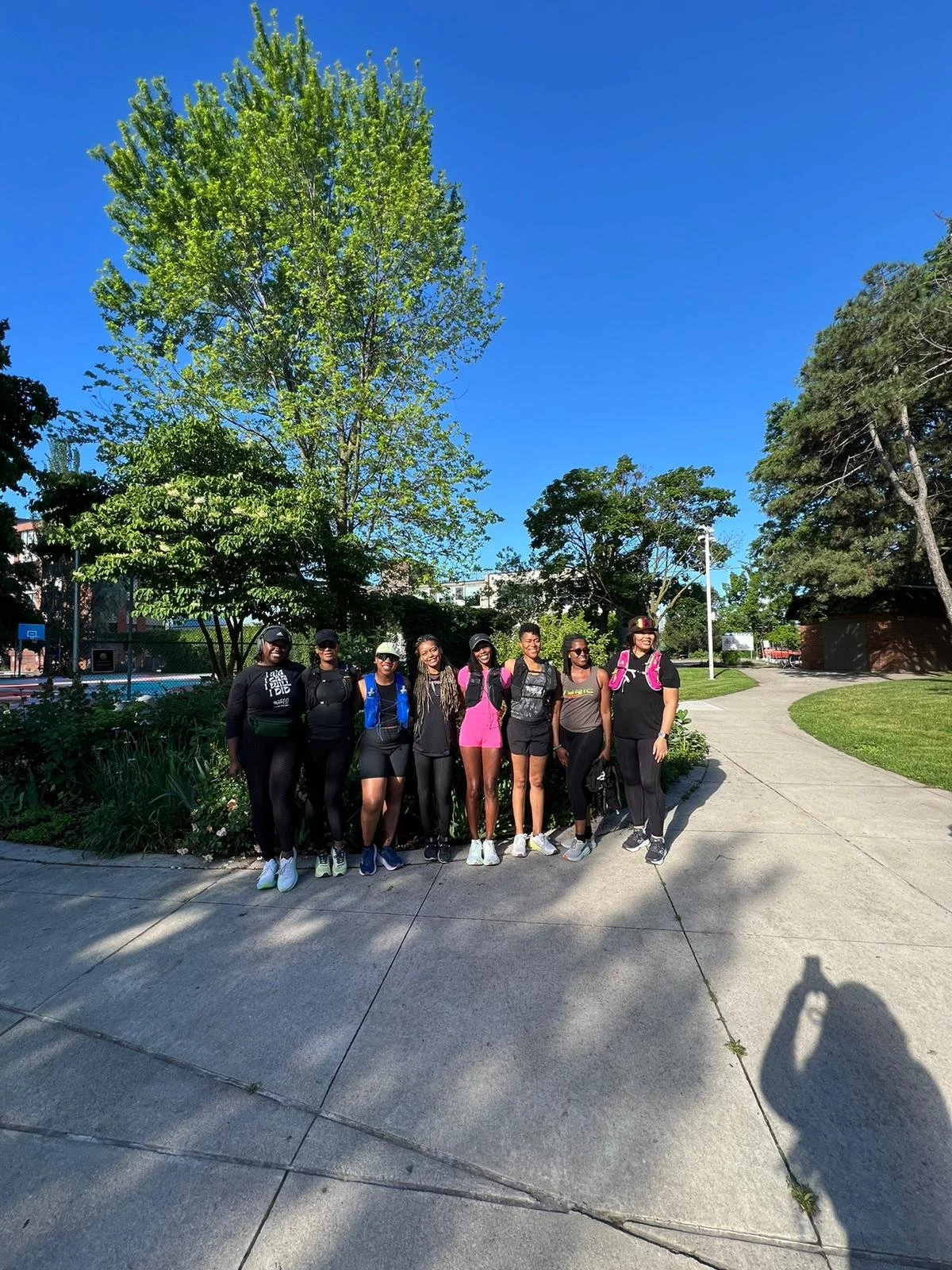 Group of eight women standing on a sidewalk in a park with trees and a blue sky in the background, dressed in athletic clothing.