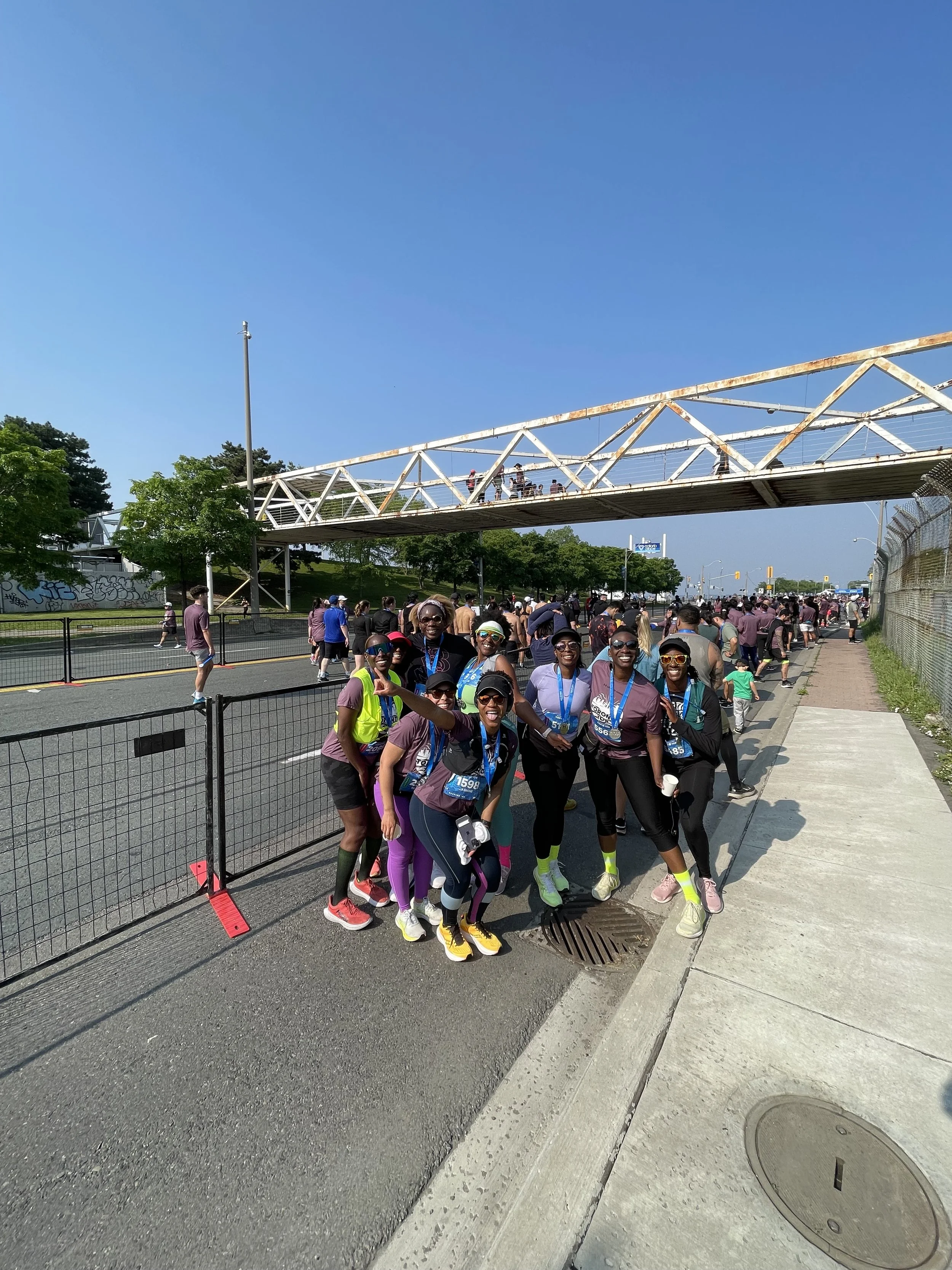 Group of runners participating in a marathon or race, posing for a photo on a city street under a pedestrian bridge, with other participants in the background.