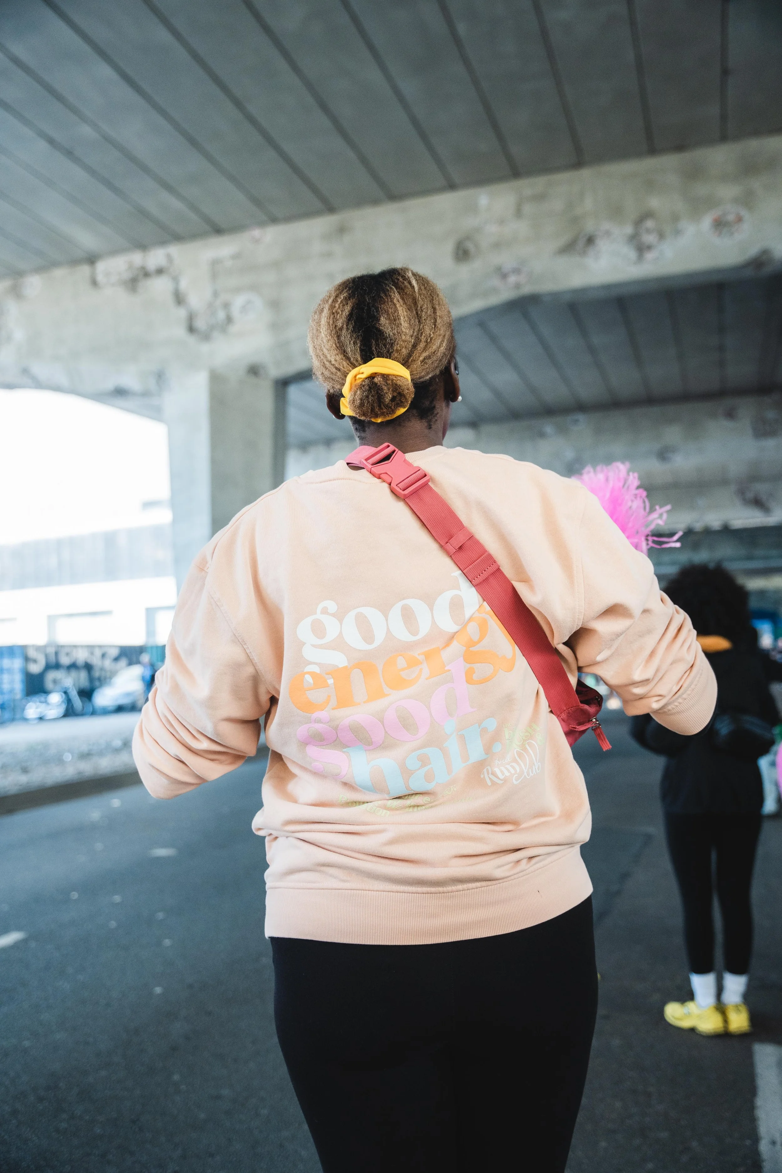 A woman with curly hair tied back with a yellow scrunchie, wearing a beige sweatshirt with multicolored text reading 'good energy good hair' and a red strap across her shoulder, standing under a bridge.