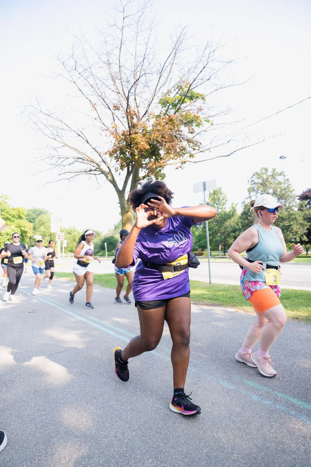 Group of women participating in a road race, with one woman in a purple shirt and black shorts in the foreground, making a hand gesture, and others running behind on a sunny day with trees and a leafless tree in the background.