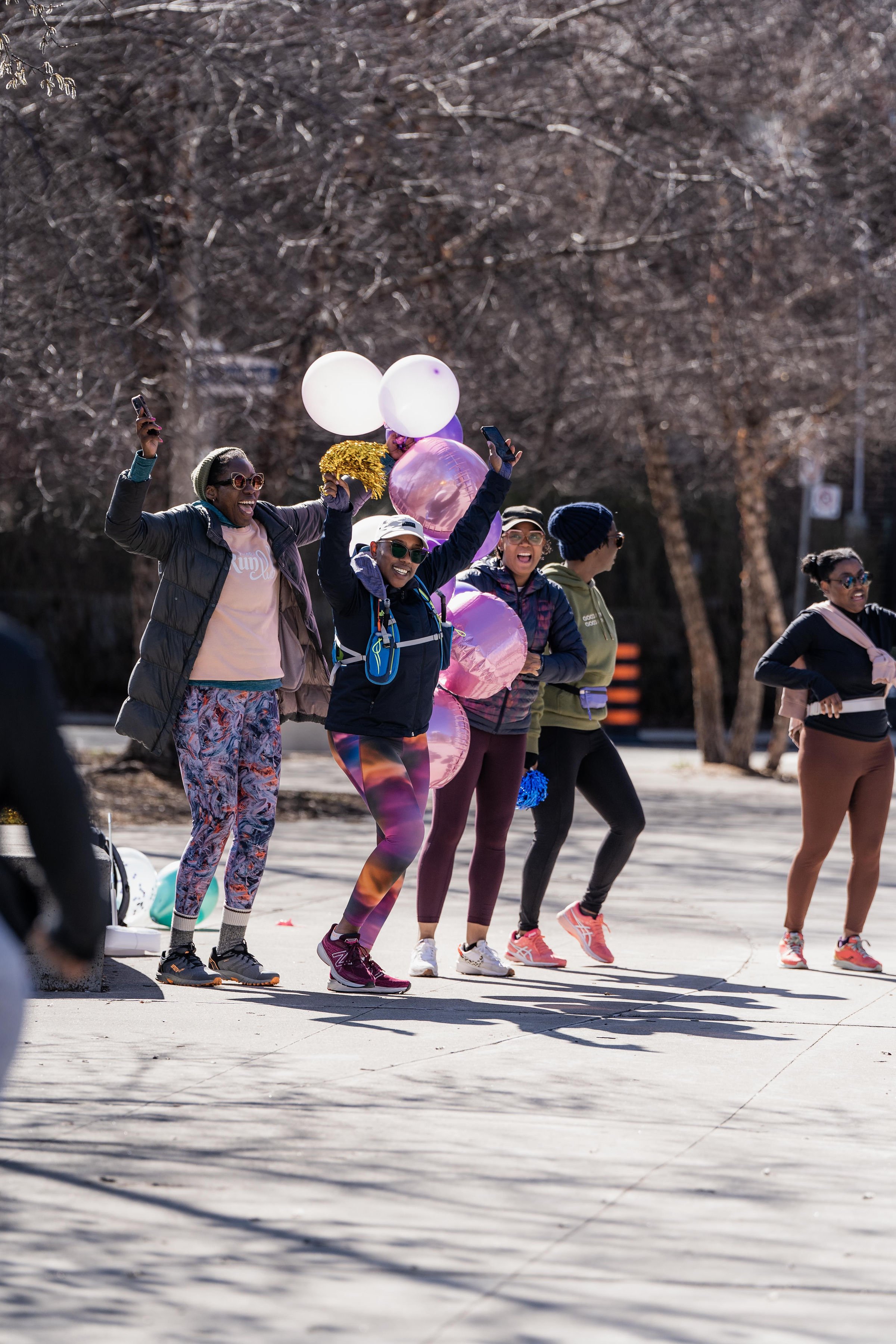 Group of women participating in outdoor fitness event, some holding balloons, dressed in activewear, on a paved path with leafless trees in the background.