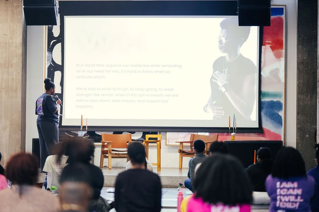 A woman presents to a seated audience in a large room with a projection screen behind her displaying a slide. The room is decorated with colorful murals and chairs are set up for the audience.