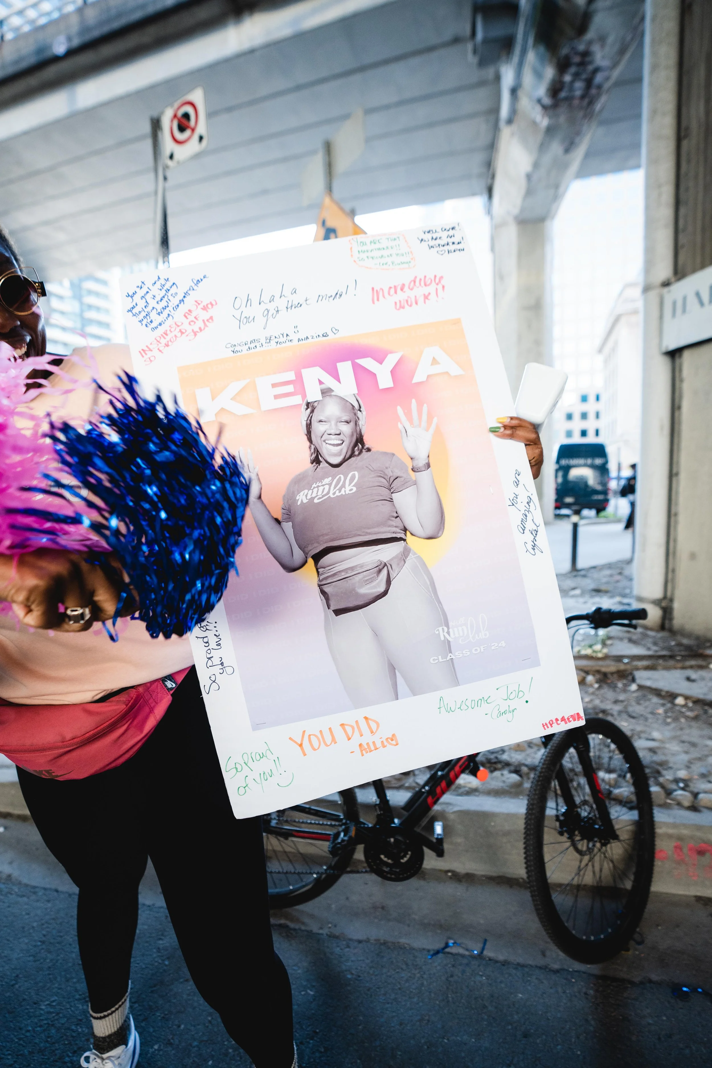 Person holding a large congratulatory poster featuring a black-and-white photo of a woman with a big smile, in an outdoor urban setting with a bicycle nearby.