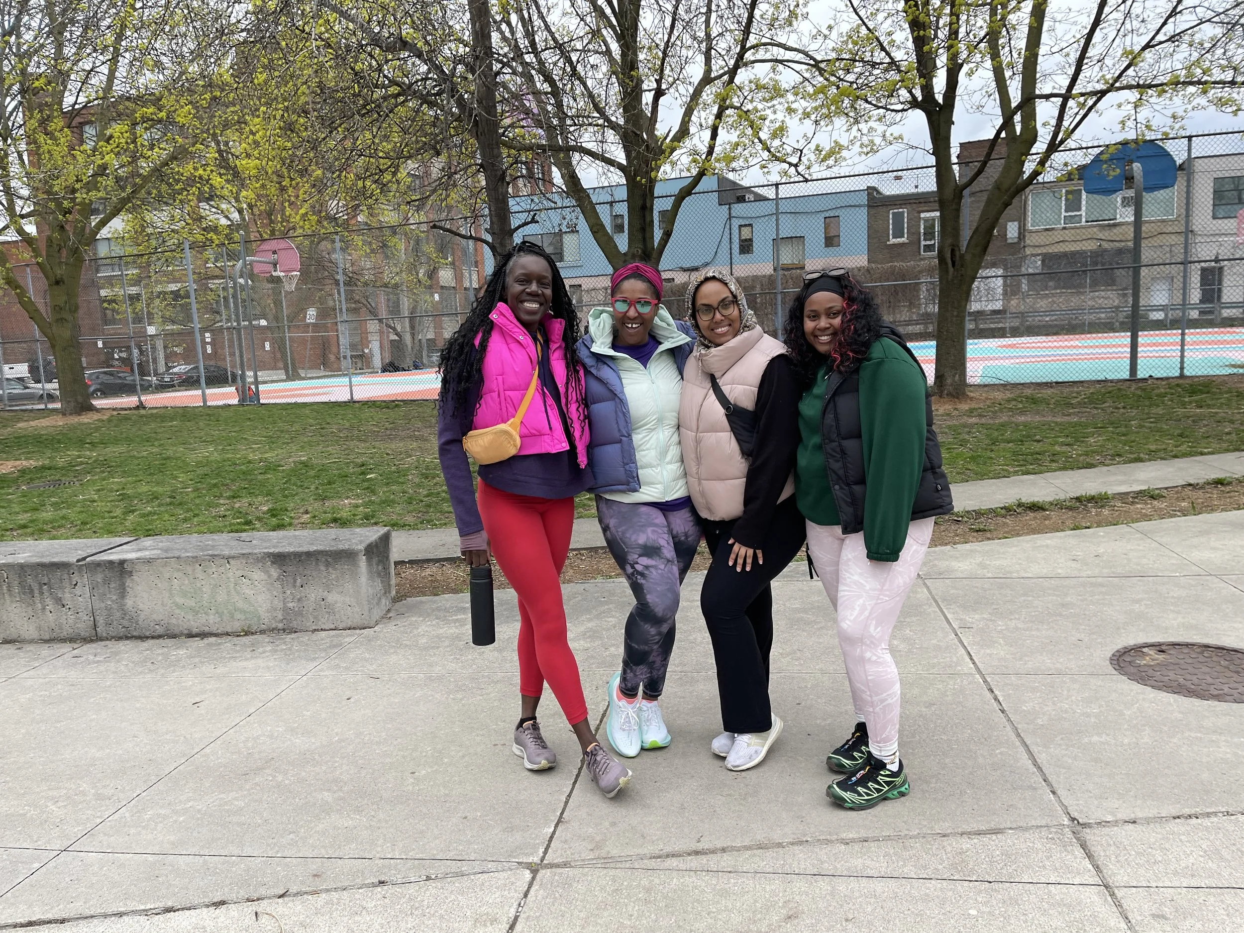 Four women standing together outdoors on a sidewalk, smiling, with trees, a chain-link fence, a basketball court, and residential buildings in the background.