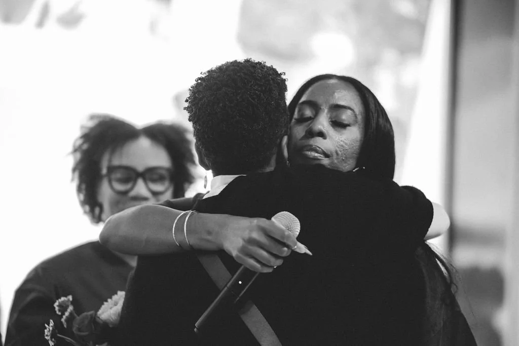 Three women engaging in an emotional embrace, one holding a microphone, in a black and white photo.
