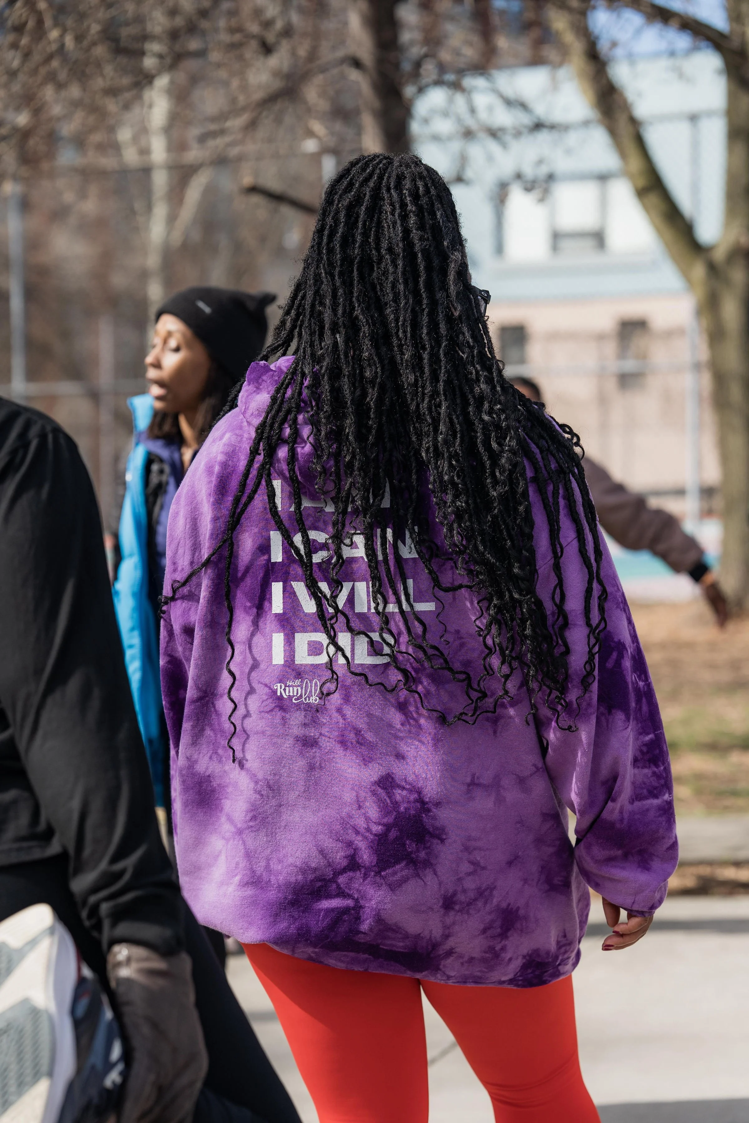 Back view of a woman with long black dreadlocks wearing a purple tie-dye hoodie and red leggings, standing outdoors on a sunny day.