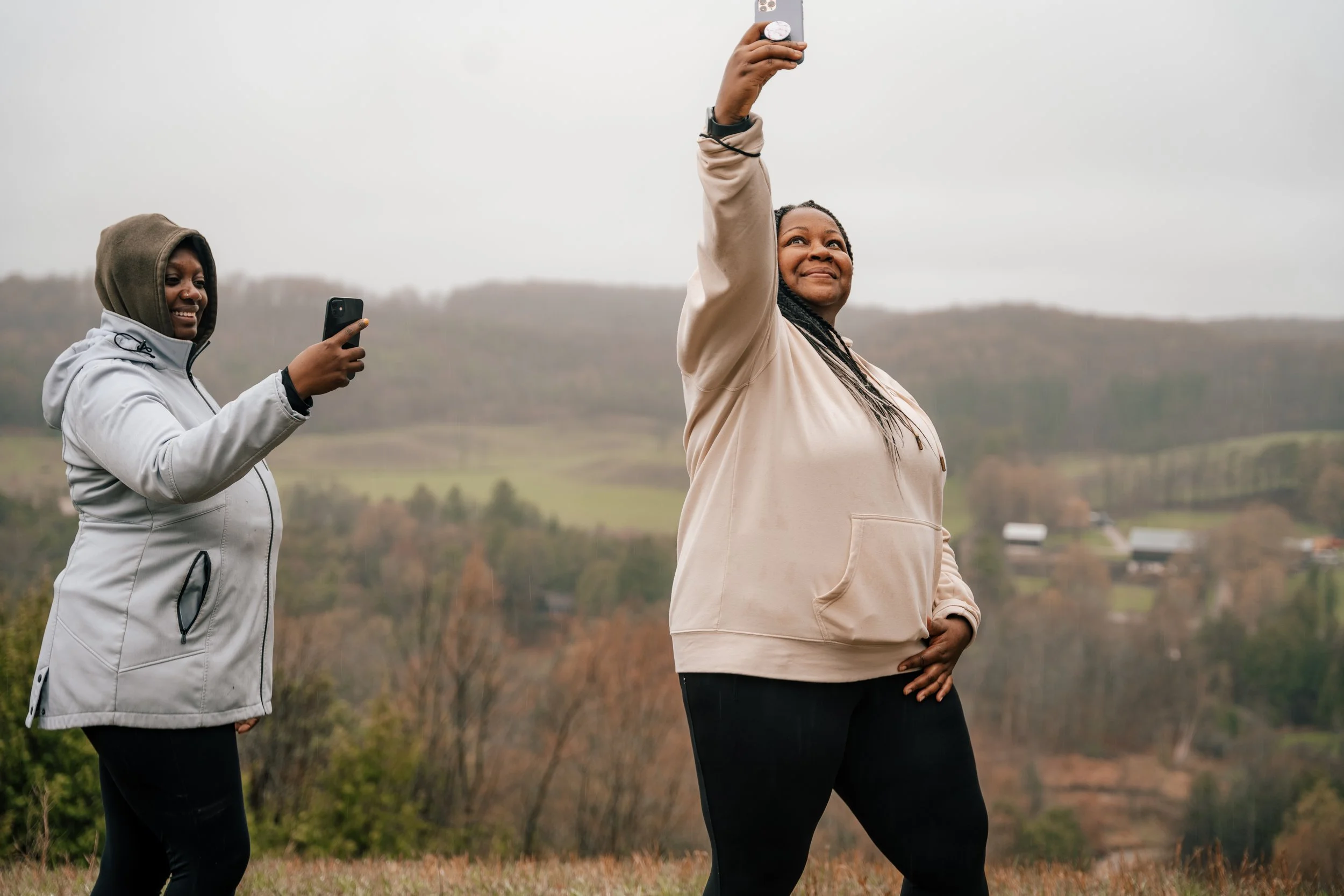 Two women outdoors taking selfies, with a scenic hilly landscape in the background.