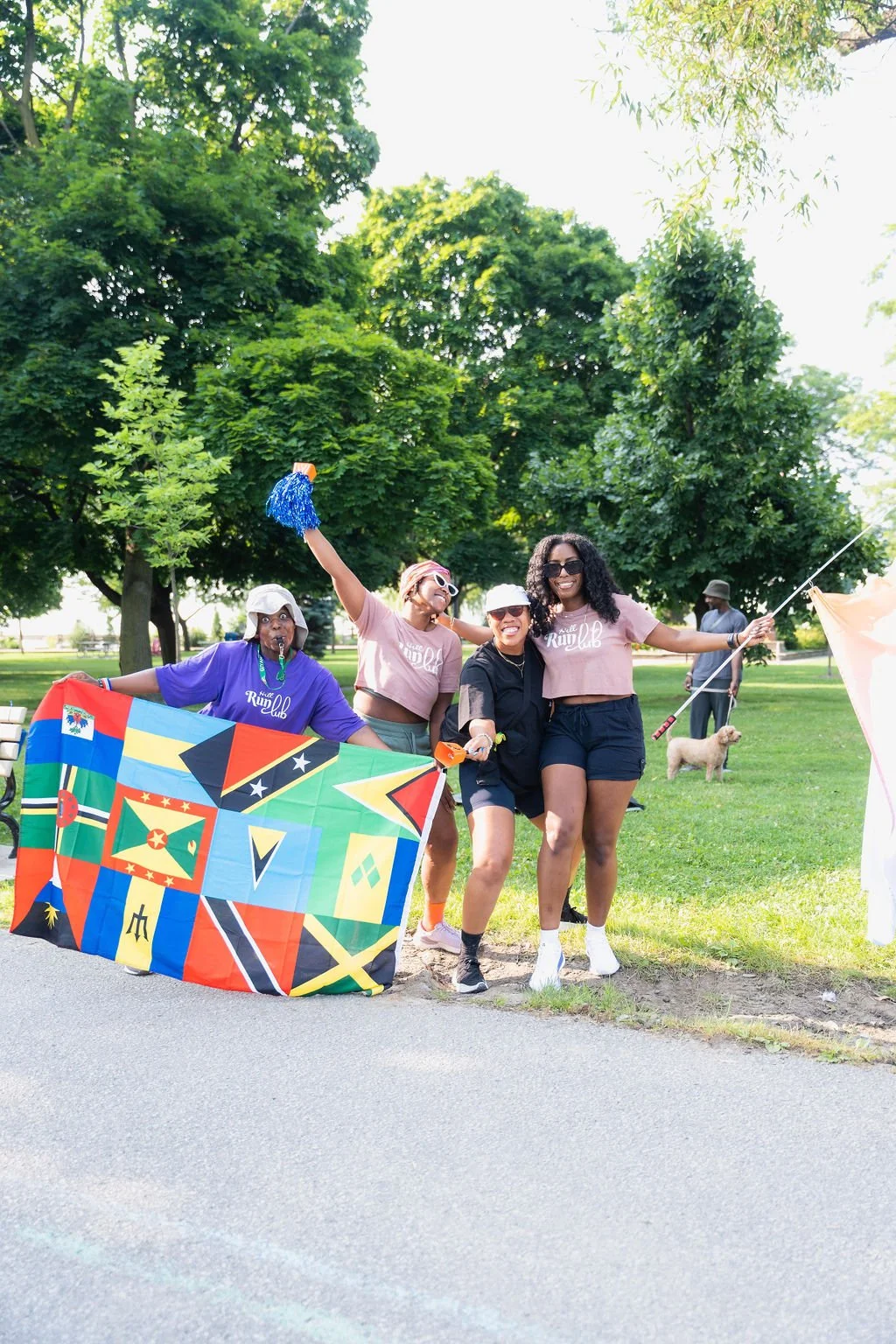 Group of four women celebrating outdoors with flags and banners on a sunny day in a park, with trees and a person walking a dog in the background.
