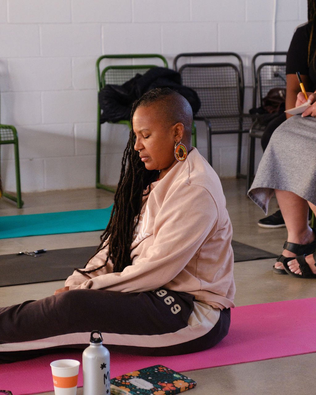 A woman meditating on a pink yoga mat in a room with chairs and a white brick wall.