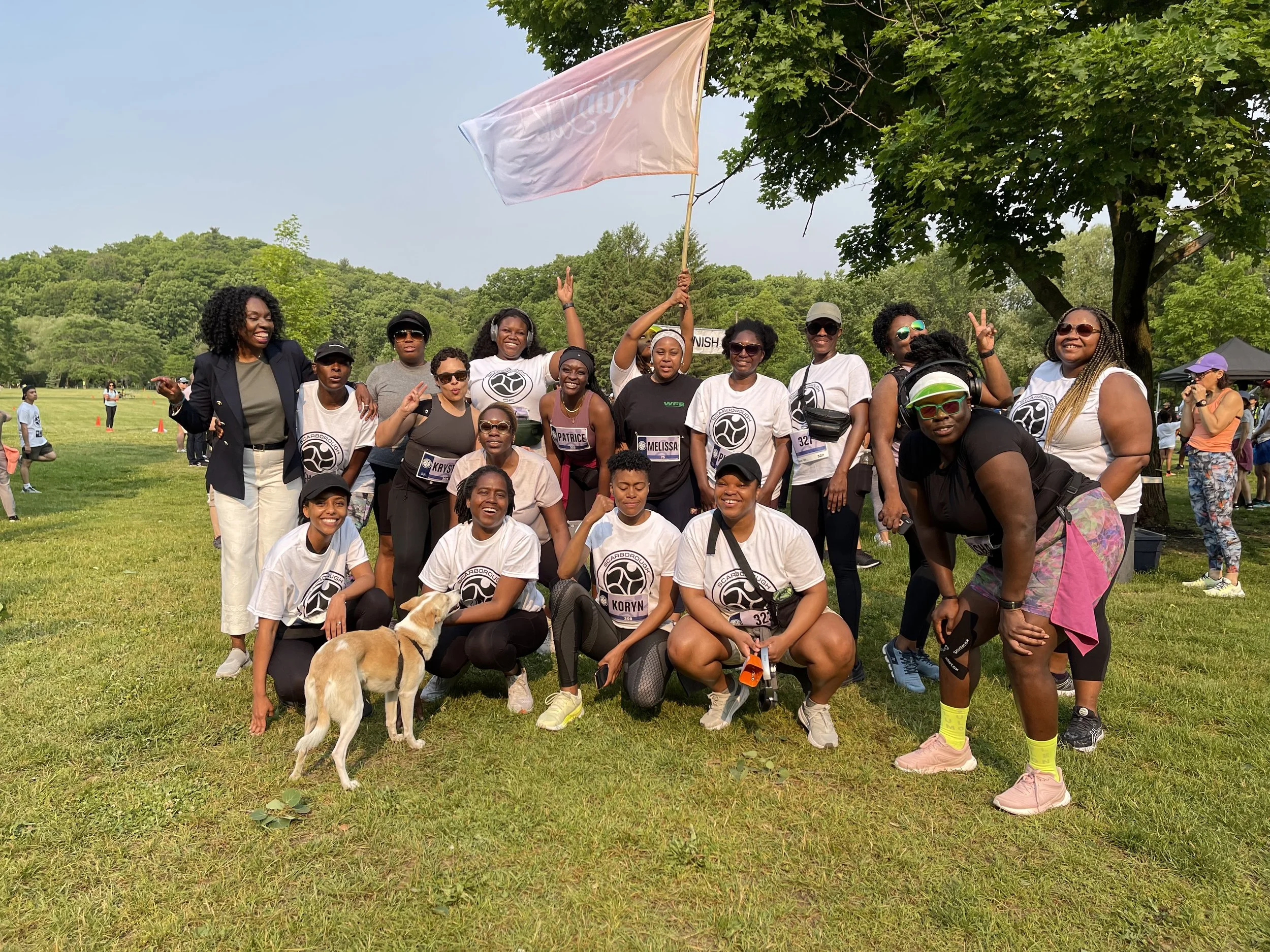 A group of people gathered outdoors in a park, posing for a photo during a running or fitness event. They are smiling, some making peace signs, and wearing athletic clothing with event badges. There is a dog in the front stage, and a person in the ba