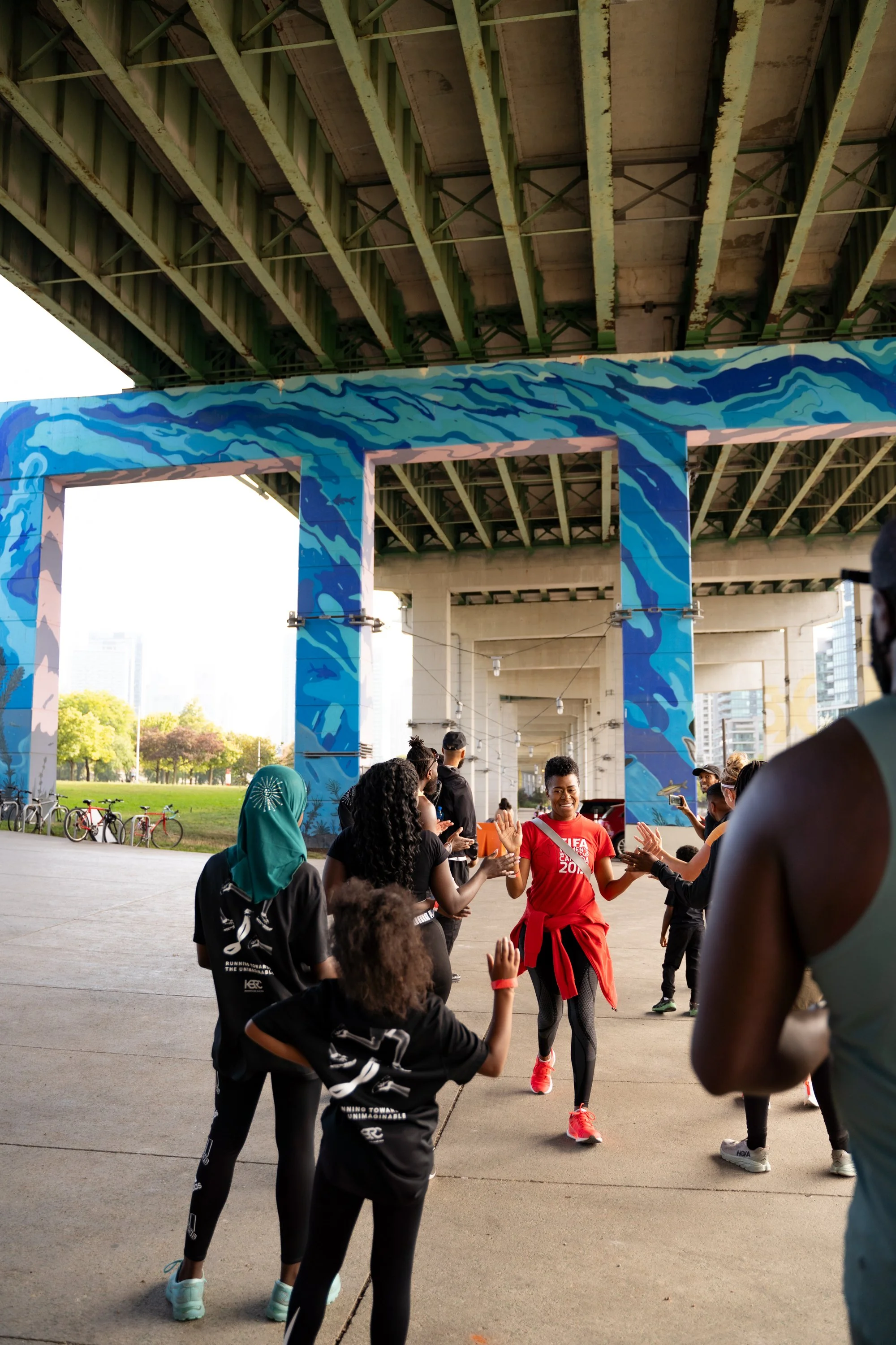 People participating in a running event under a bridge, with some high-fiving each other. The bridge has blue and teal graffiti art, and bicycles are seen parked nearby in a park with greenery.