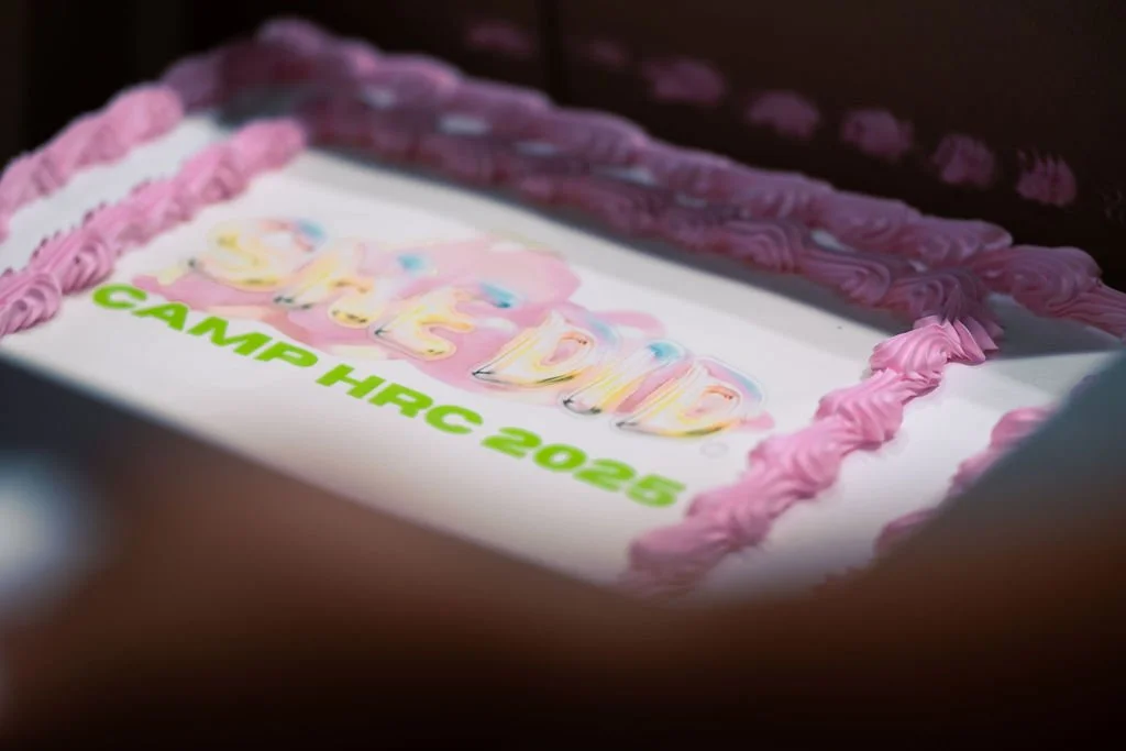 Cake with pink frosting border and a white top, decorated with colorful text that says "Camp Dads" and "Camp Hrc 2025" in green, partially in shadow.