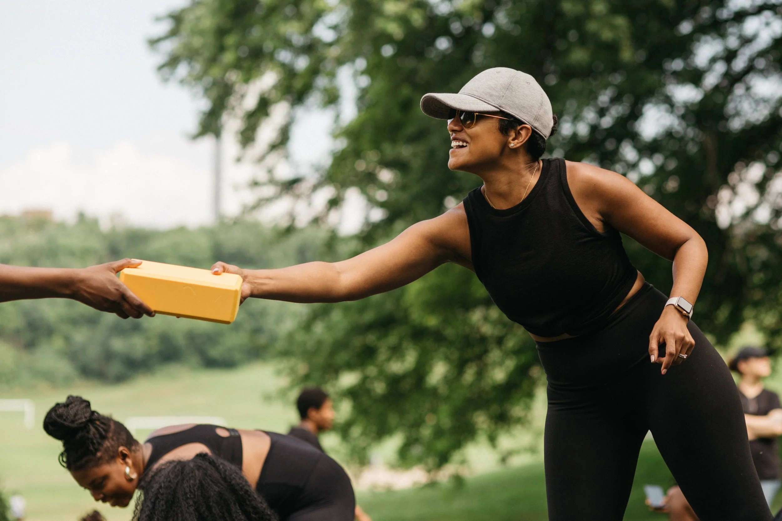 A woman in athletic clothing and a baseball cap is handing a yellow foam block to someone outside.