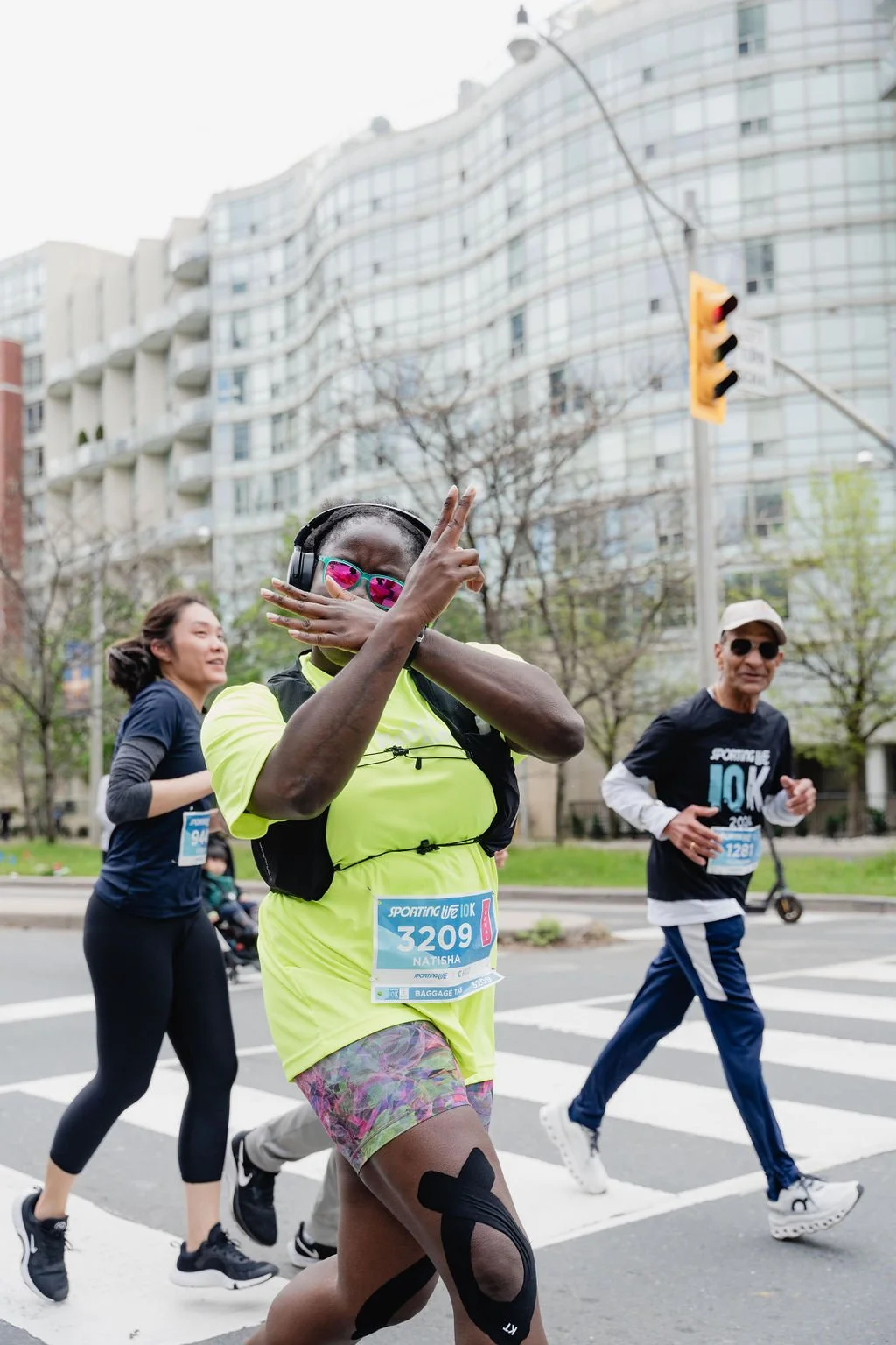 Group of runners participating in a marathon or race on city streets, with one woman in bright yellow shirt making a peace sign gesture.