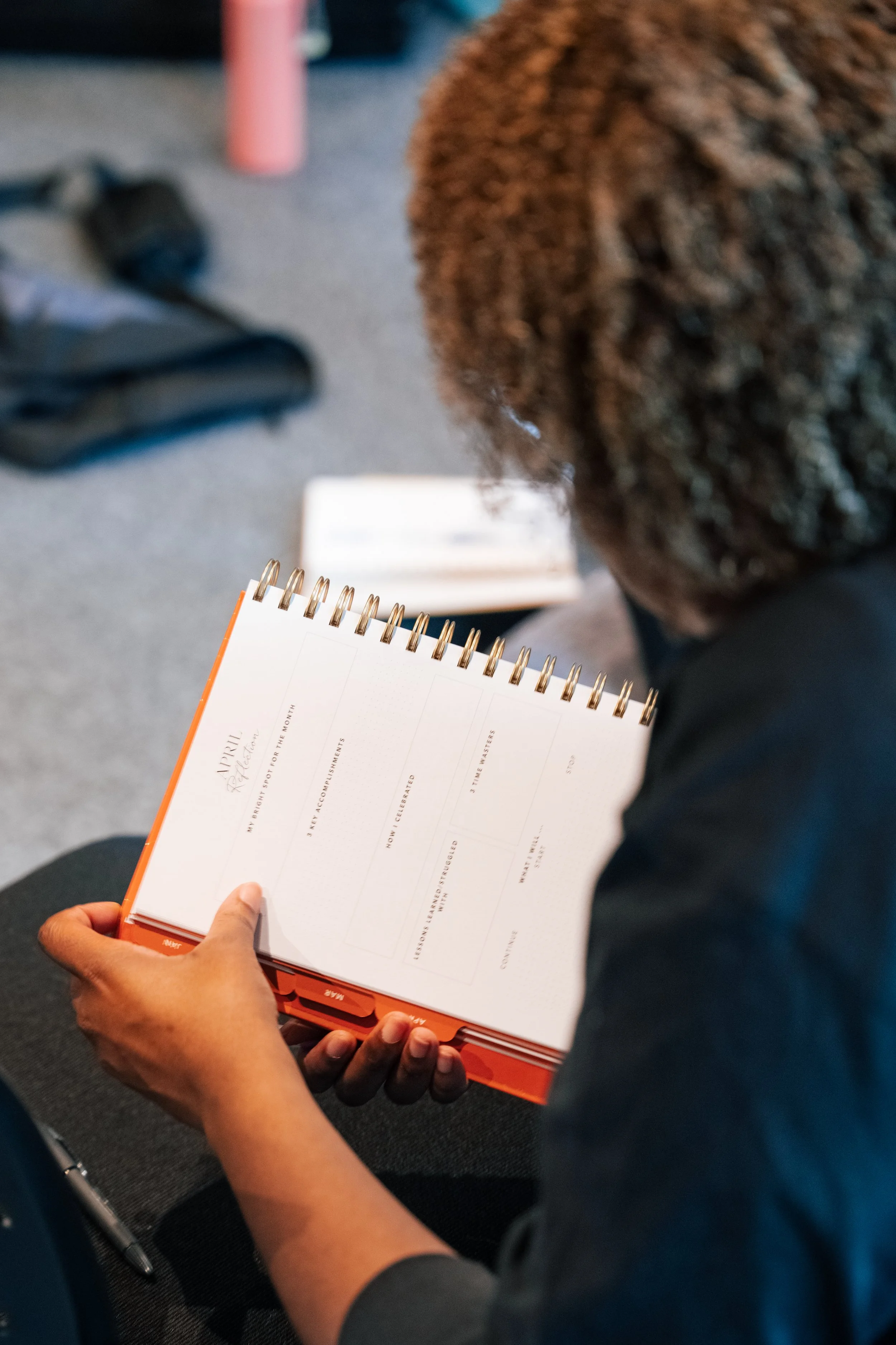 Person sitting and looking at a planner or journal, with a pen on their lap. The planner is open to a page titled 'April' with lined sections for notes and planning.
