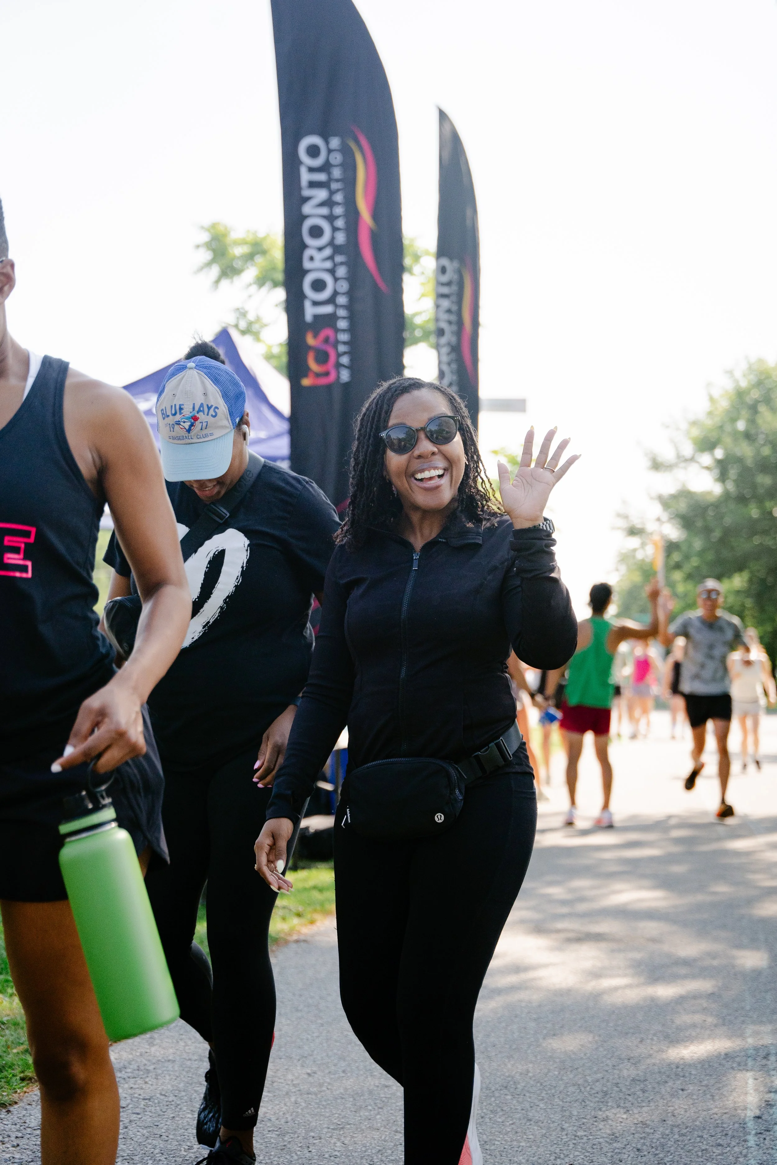 A woman wearing sunglasses and black athletic wear is smiling and waving at a community walk event with others in the background.