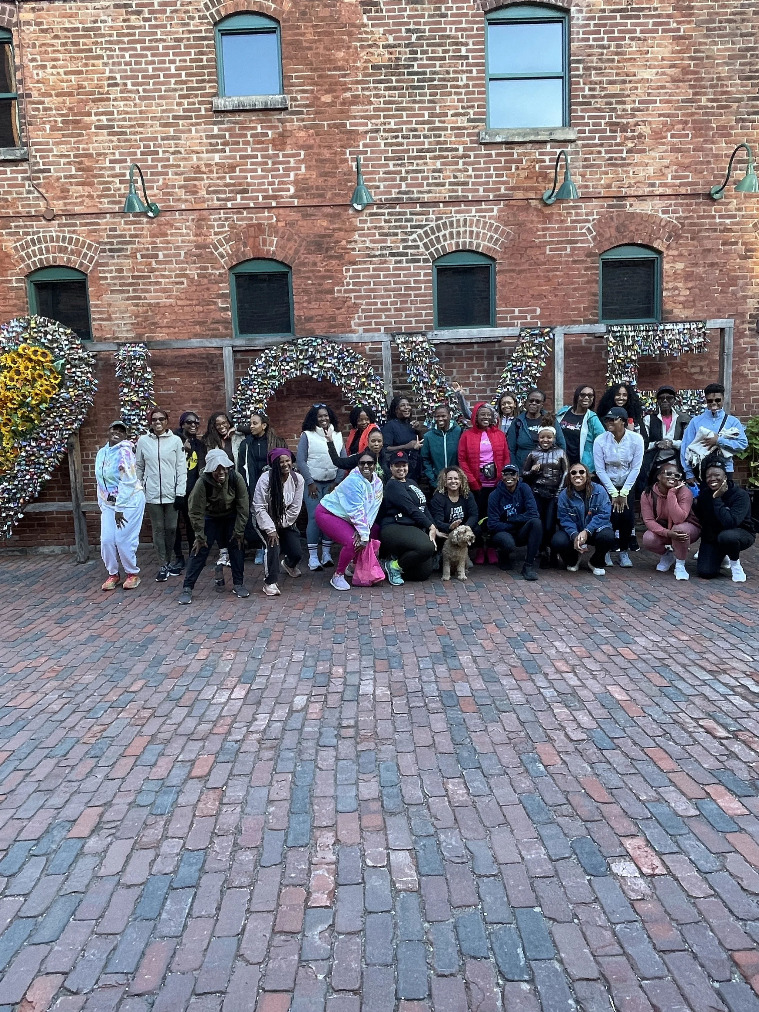 A group of people posing for a photo in front of a large decorative sign that reads 'LOVE' made of numerous small items, in an outdoor brick-paved area, with a brick building in the background.