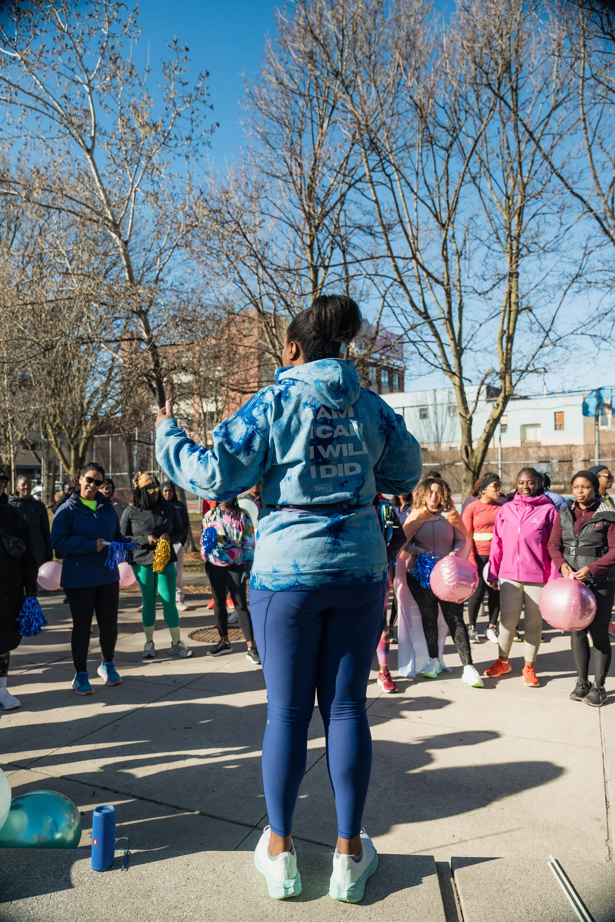 A group of women gathered outdoors on a sunny day, some holding pink and blue balloons, in what appears to be a community event or walk, with leafless trees and buildings in the background.
