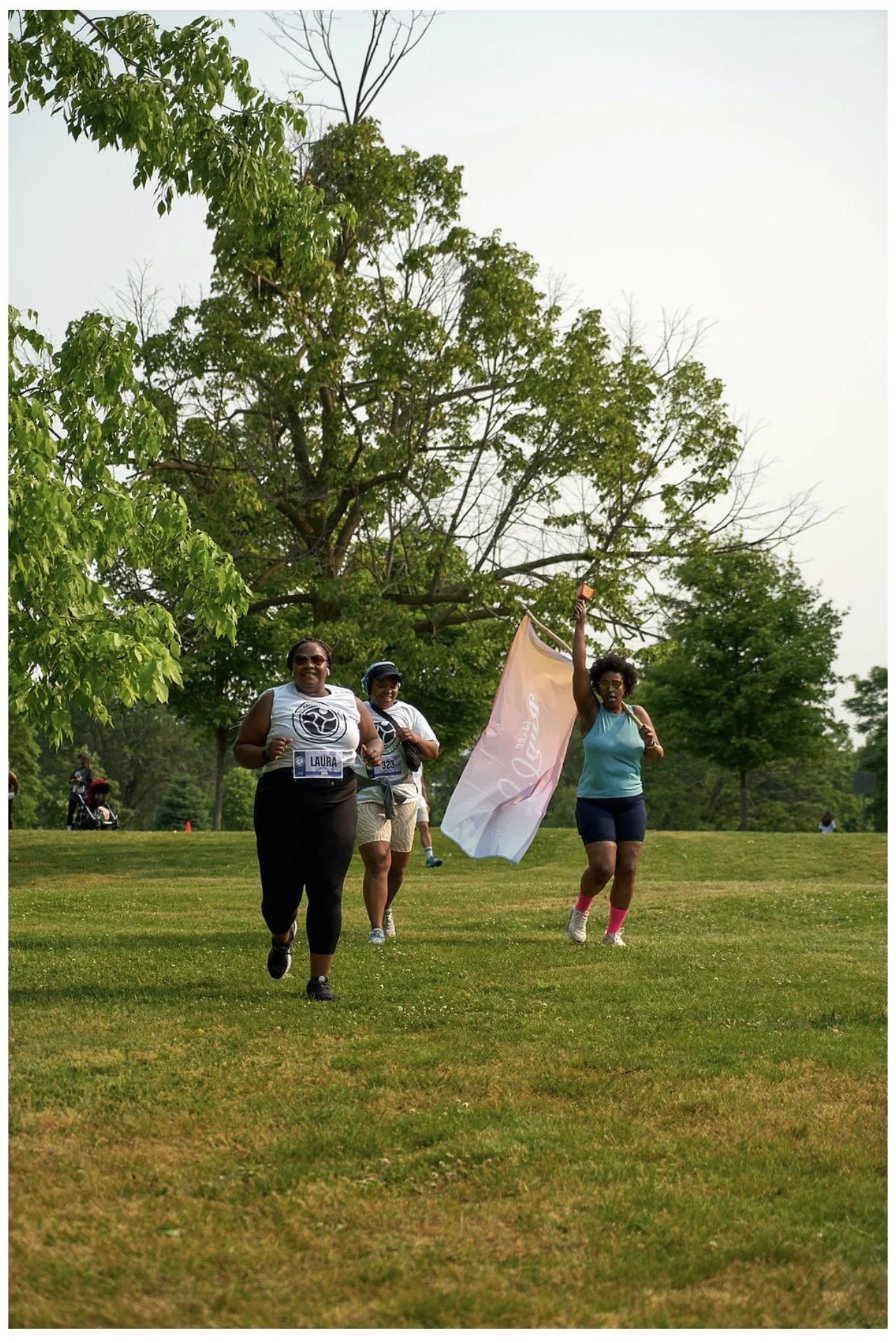 Three women running outdoors on a grassy field, with one woman carrying a flag, trees in the background, on a clear day.