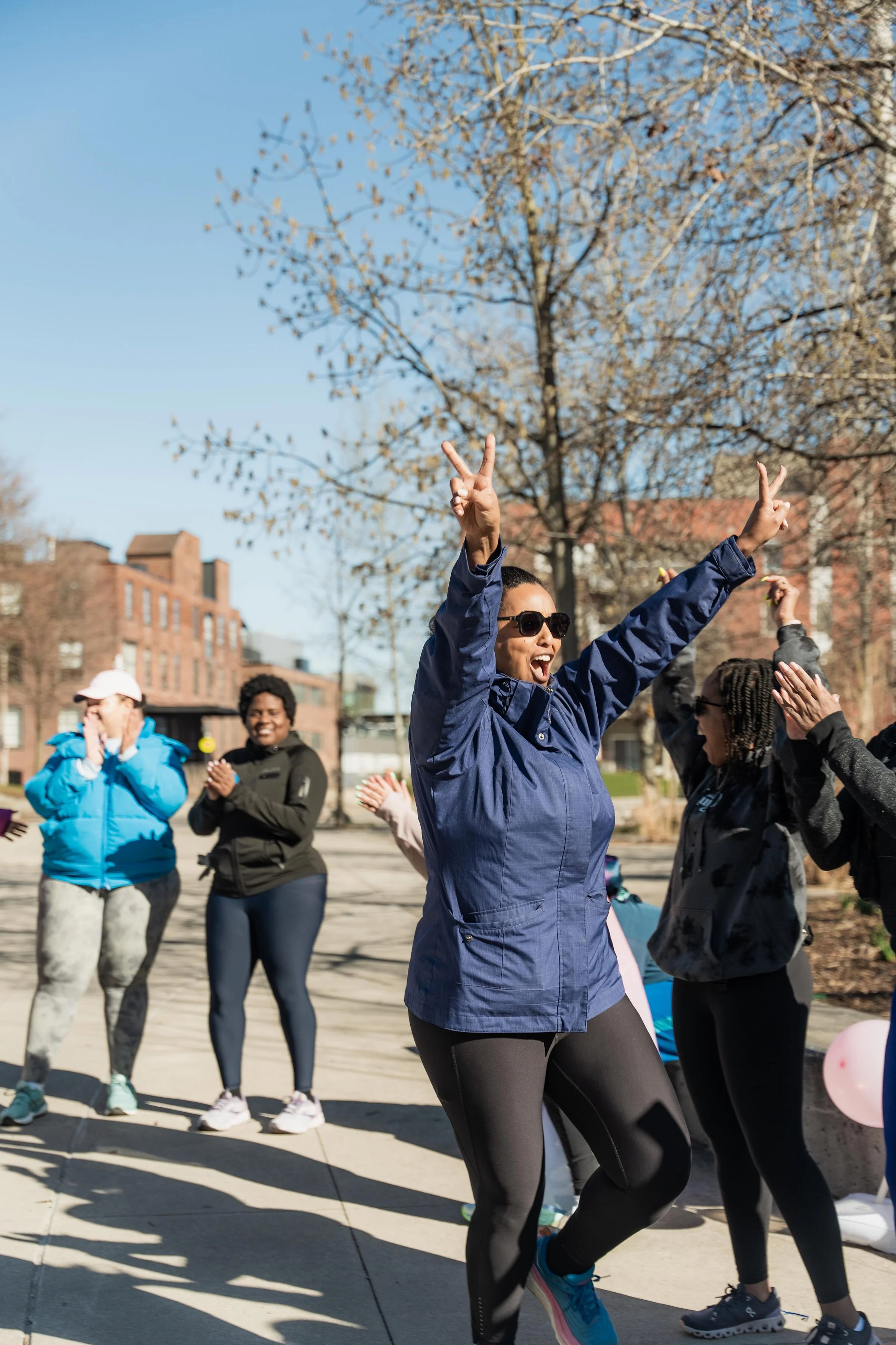 A woman wearing sunglasses and a navy jacket dancing with arms raised, surrounded by smiling people outdoors on a sunny day.