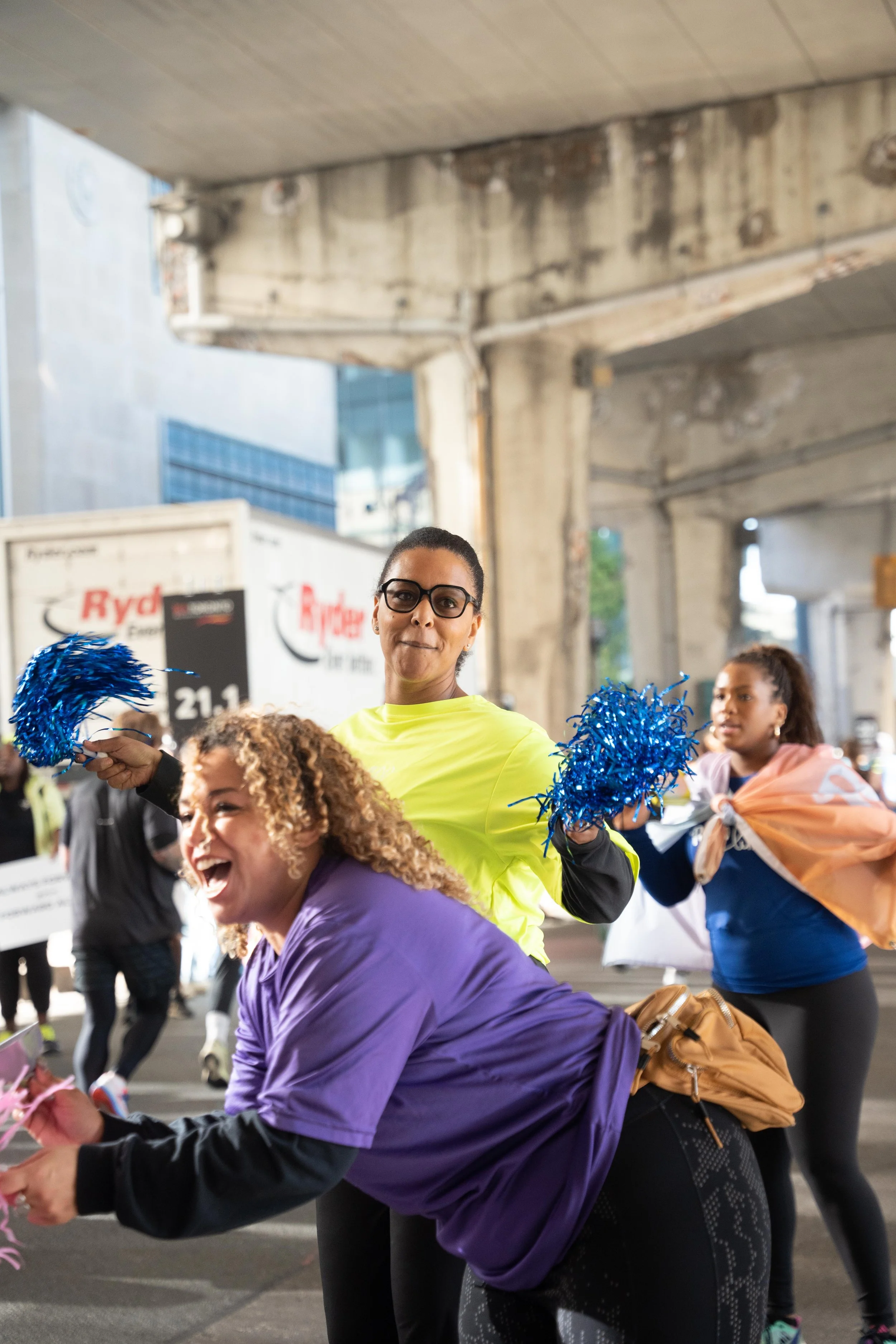 Women waving blue pom-poms and wearing colorful outfits at an outdoor event under a concrete structure.