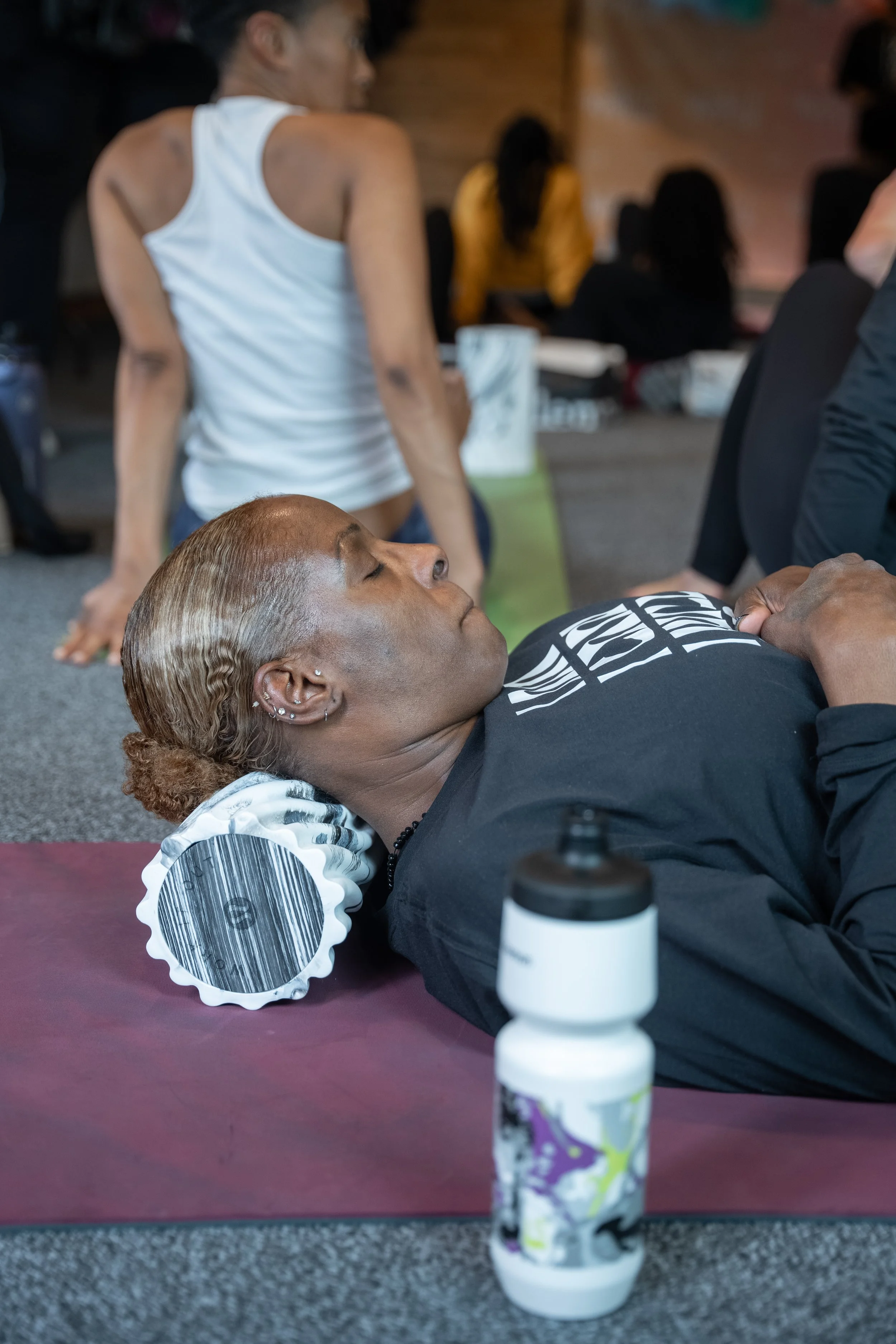 A woman with dyed hair lying on a yoga mat with a foam roller under her neck, in a gym or fitness studio surrounded by other people.
